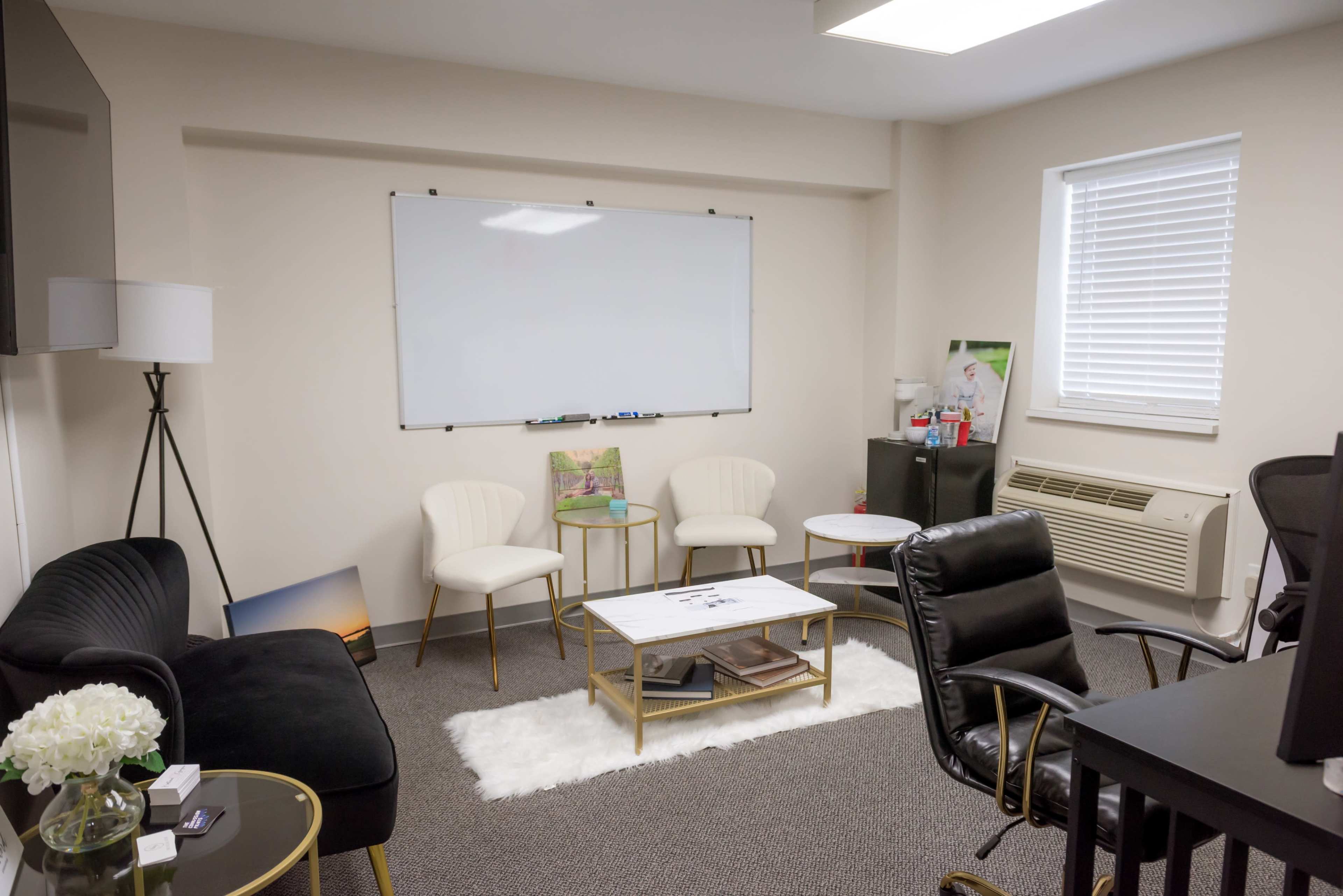 The image shows a small office space with a black couch, three white chairs, a coffee table on a rug, a black office chair, and a whiteboard on the wall.