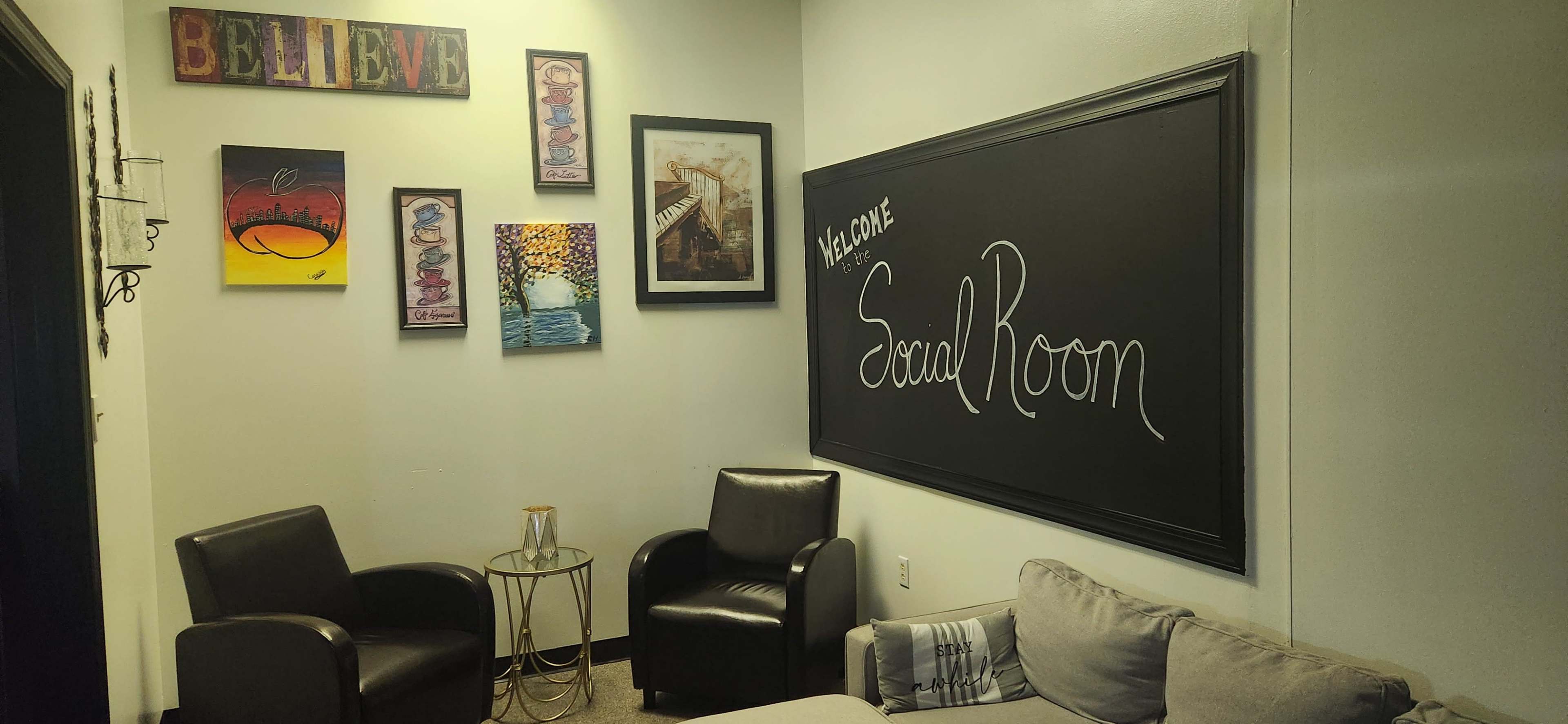 The image shows a cozy room labeled "Social Room" with two black chairs, a couch, a small table, and various art pieces on the walls.