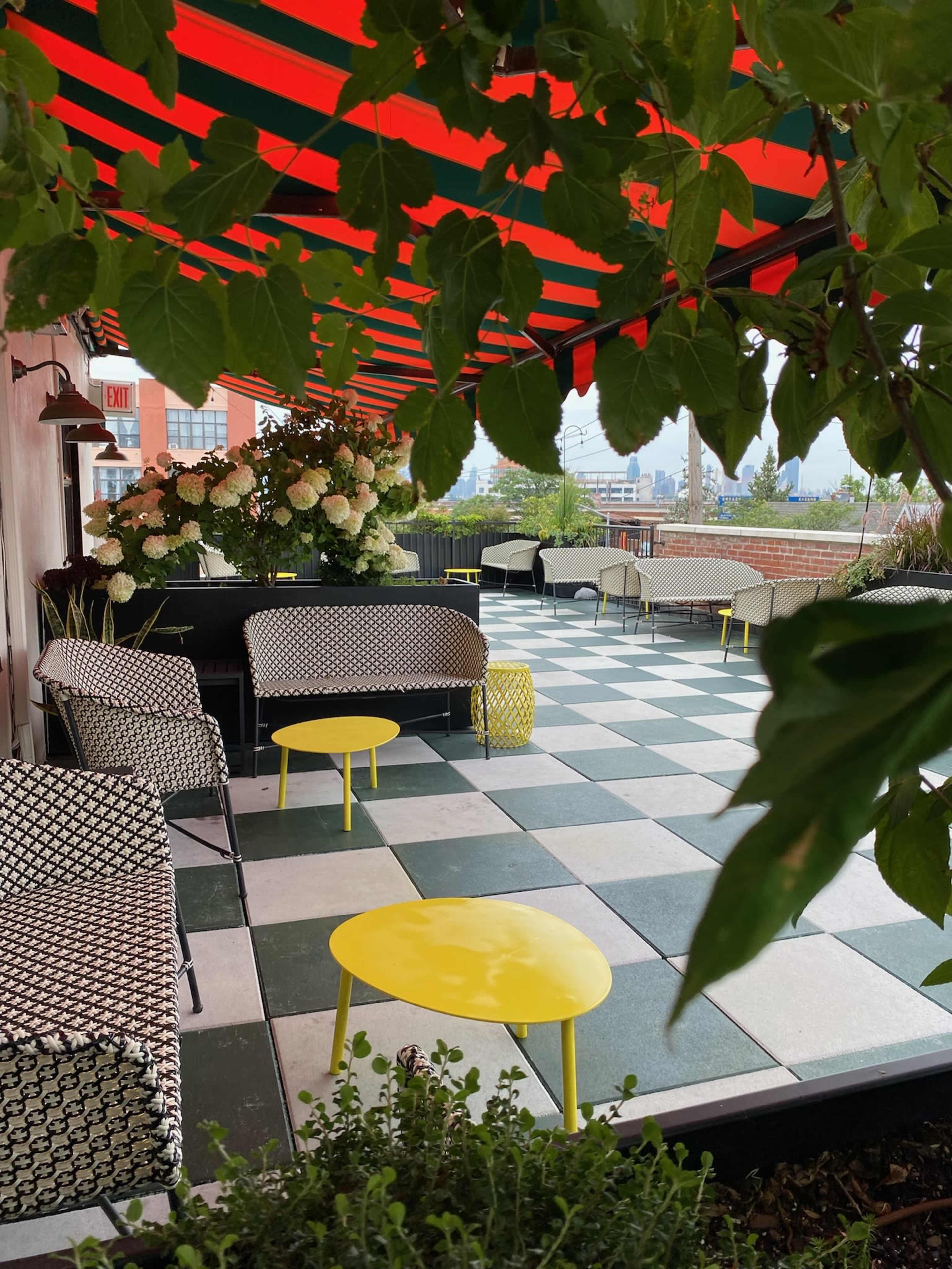 A rooftop patio features patterned seating, colorful tables, and a canopy of striped fabric with views of a city skyline.
