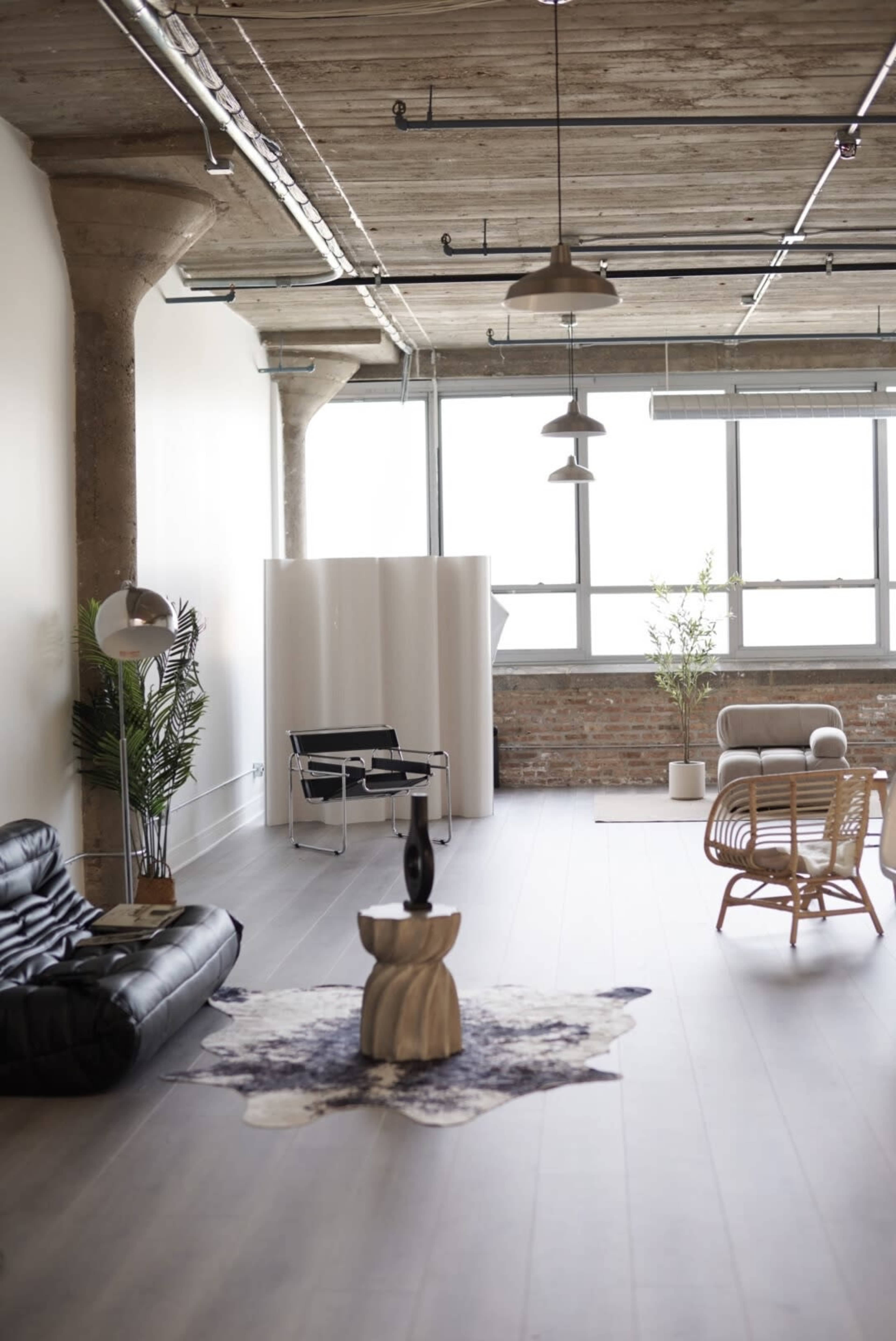 The image shows a spacious, modern loft-style room with large windows, concrete columns, and a mix of contemporary furniture including a black sofa, a light-colored armchair, and a decorative rug on the floor.