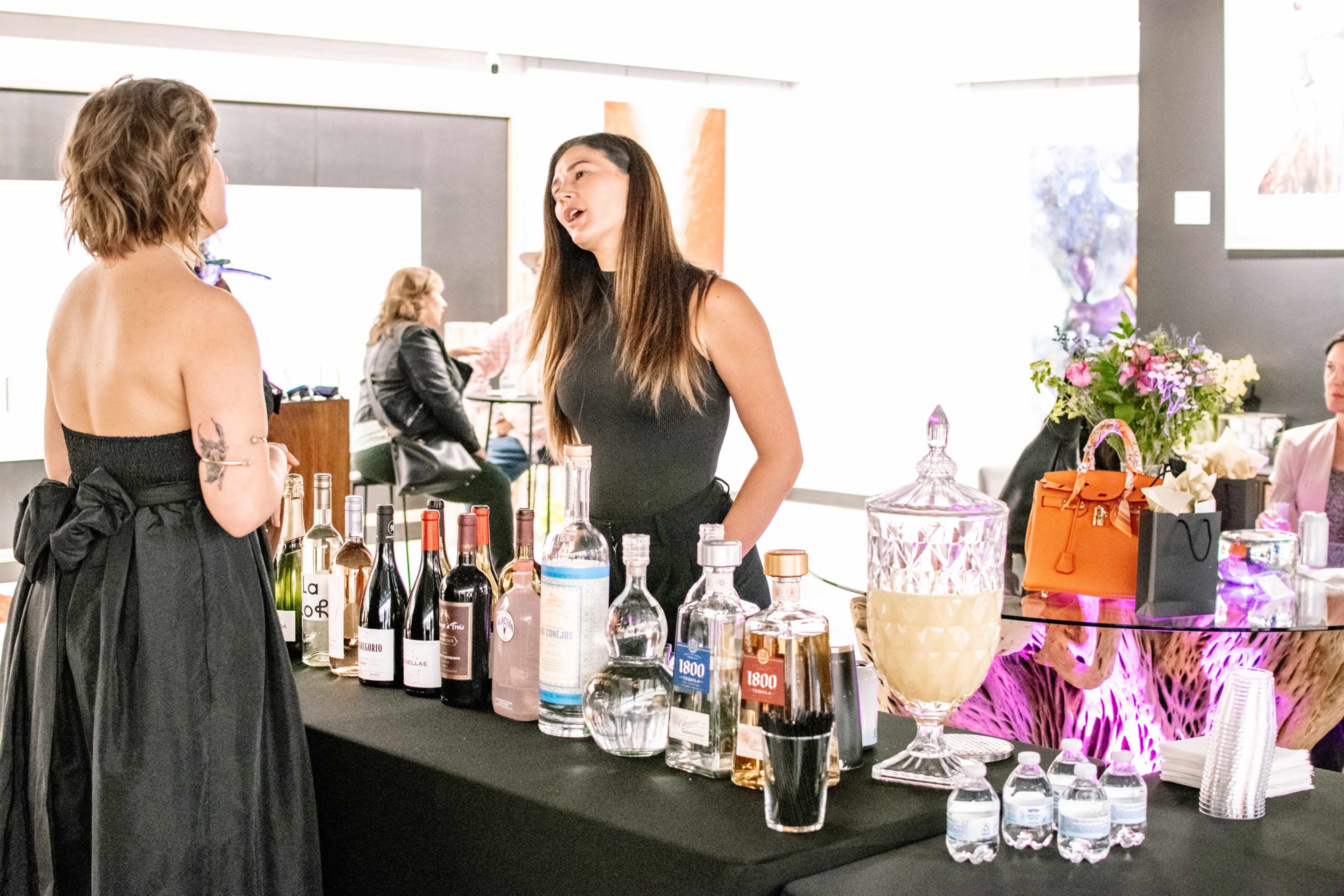 Two women converse at an elegant bar setup featuring a variety of alcoholic beverages and a decorative display.