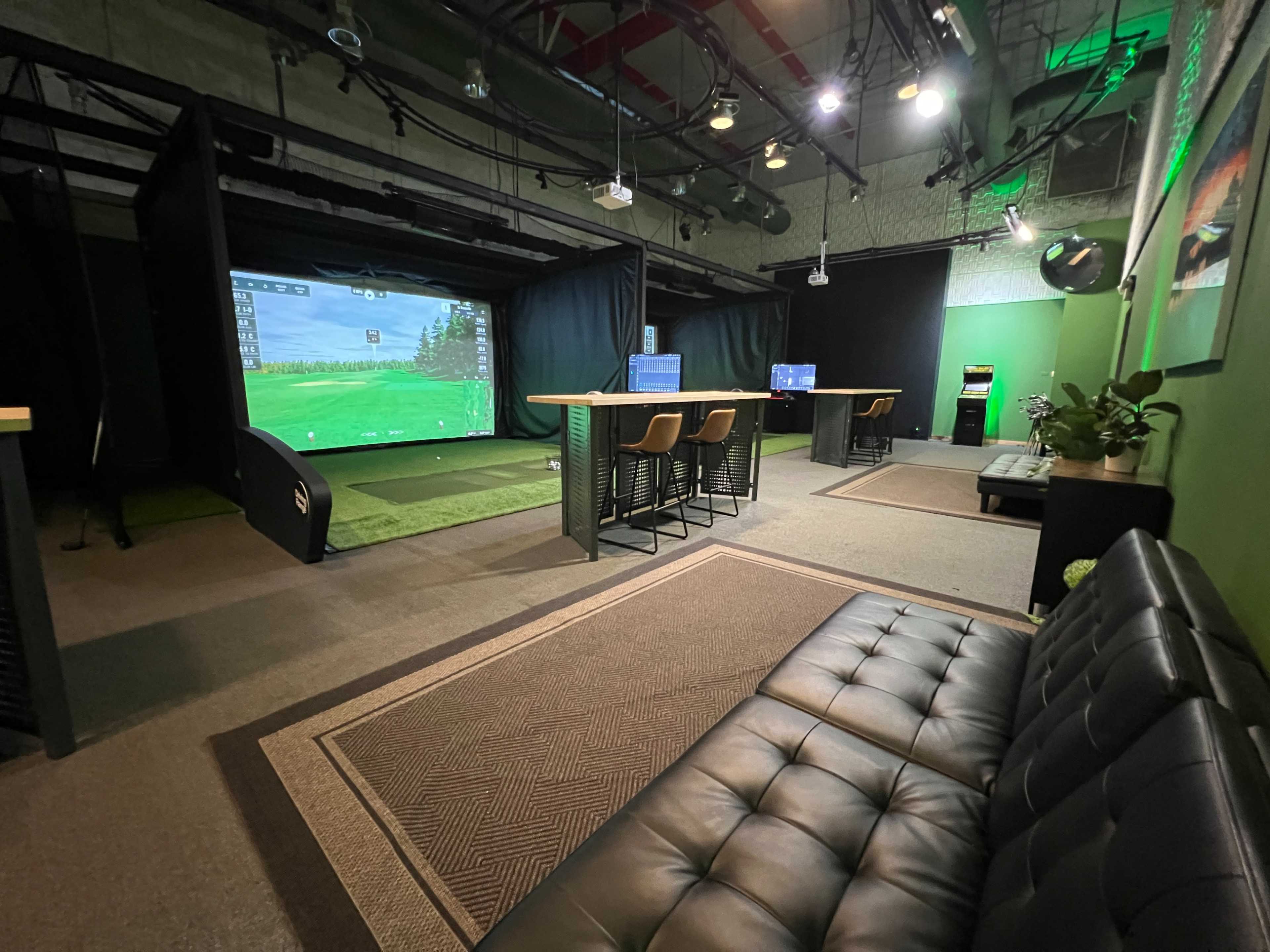 The image shows an indoor golf simulation area with a large screen displaying a golf course, surrounded by green carpeting and seating areas.