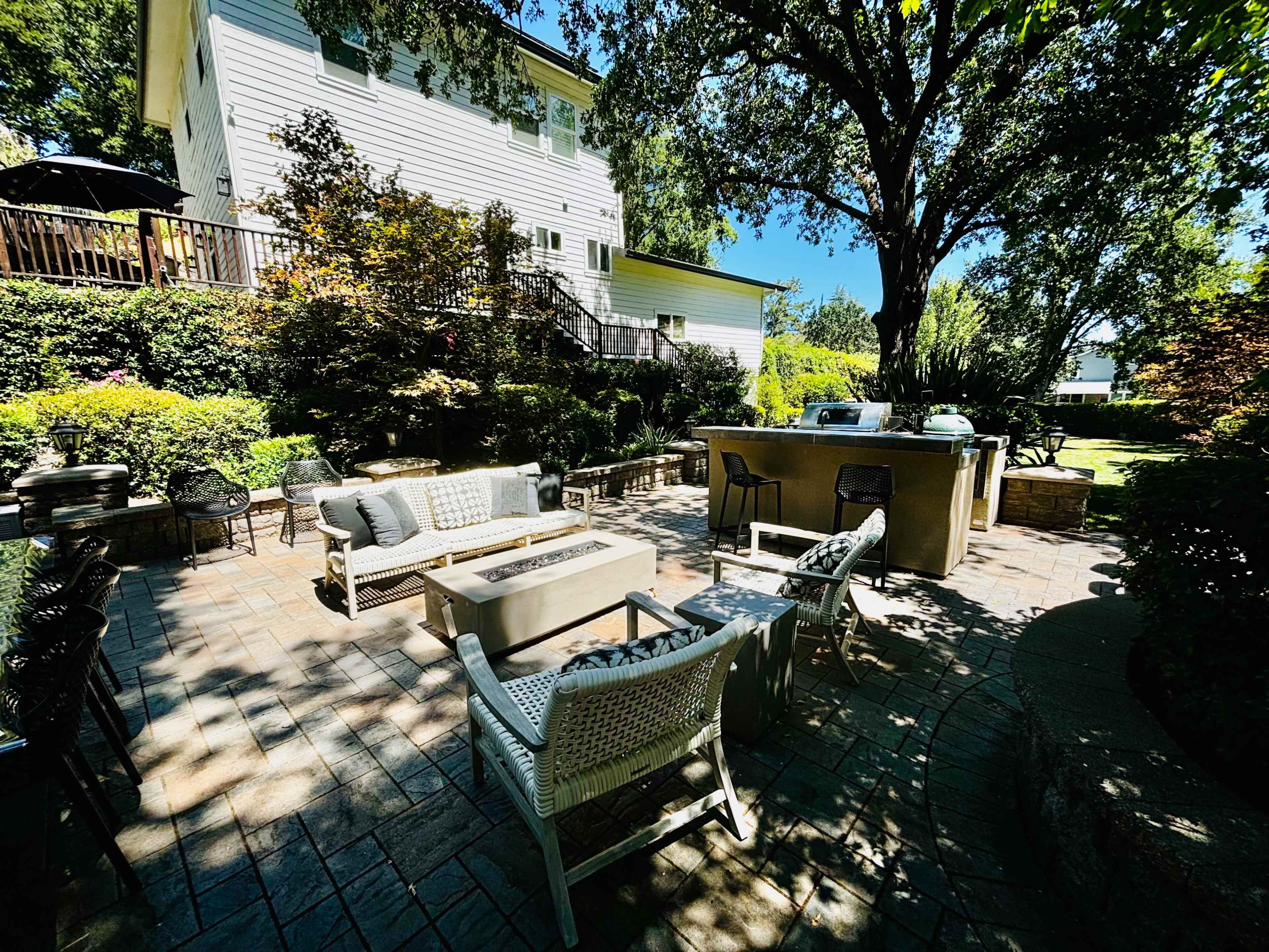 A spacious backyard patio features a set of outdoor furniture, a fire pit, and a small kitchen area, surrounded by lush greenery and trees.