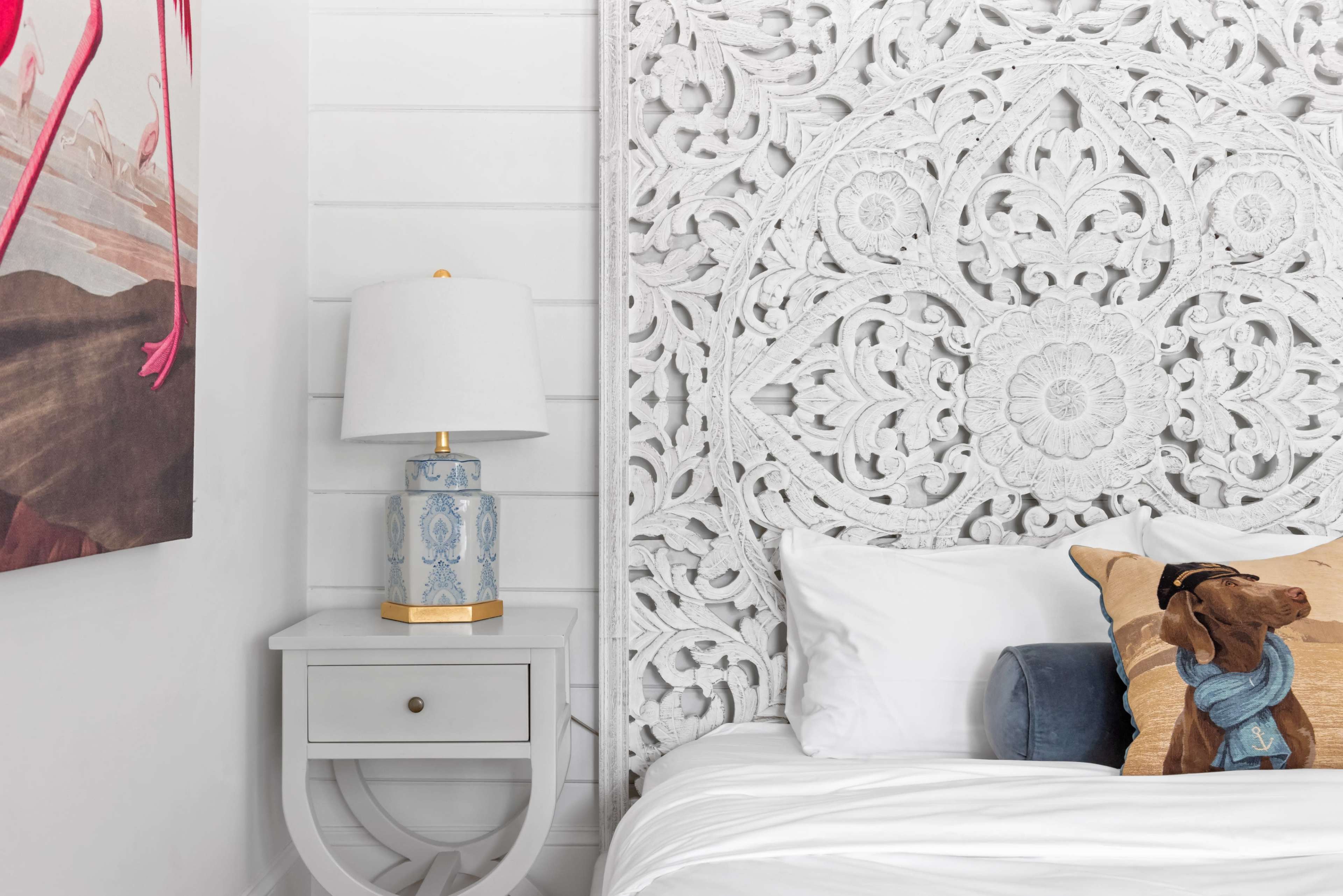 The image depicts a modern bedroom featuring a white carved wooden headboard, a bedside table with a lamp, and decorative pillows on the bed.