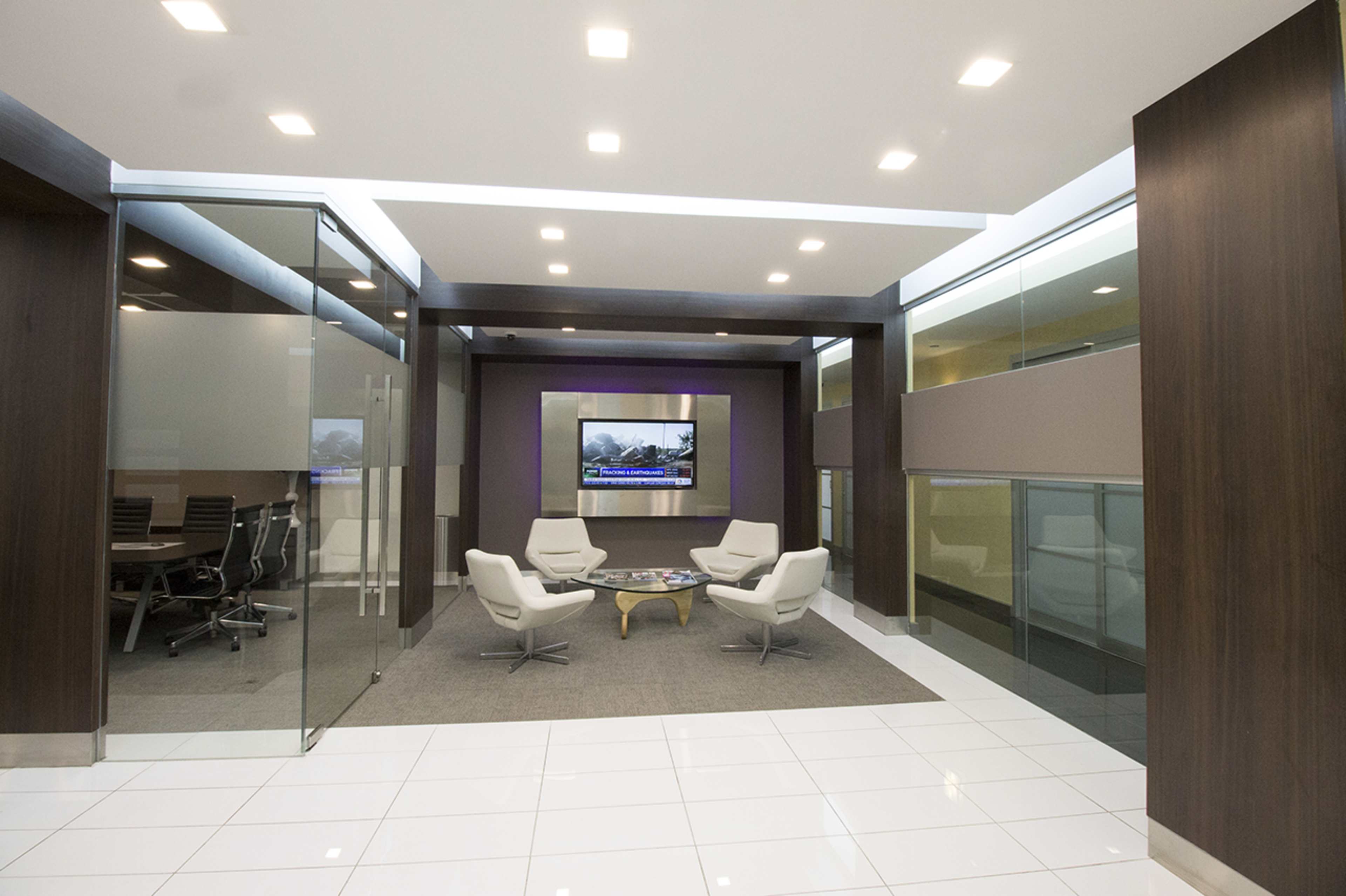 A modern office lobby with glass walls, a seating area featuring white chairs and a round coffee table, and a wall-mounted television displaying a landscape.