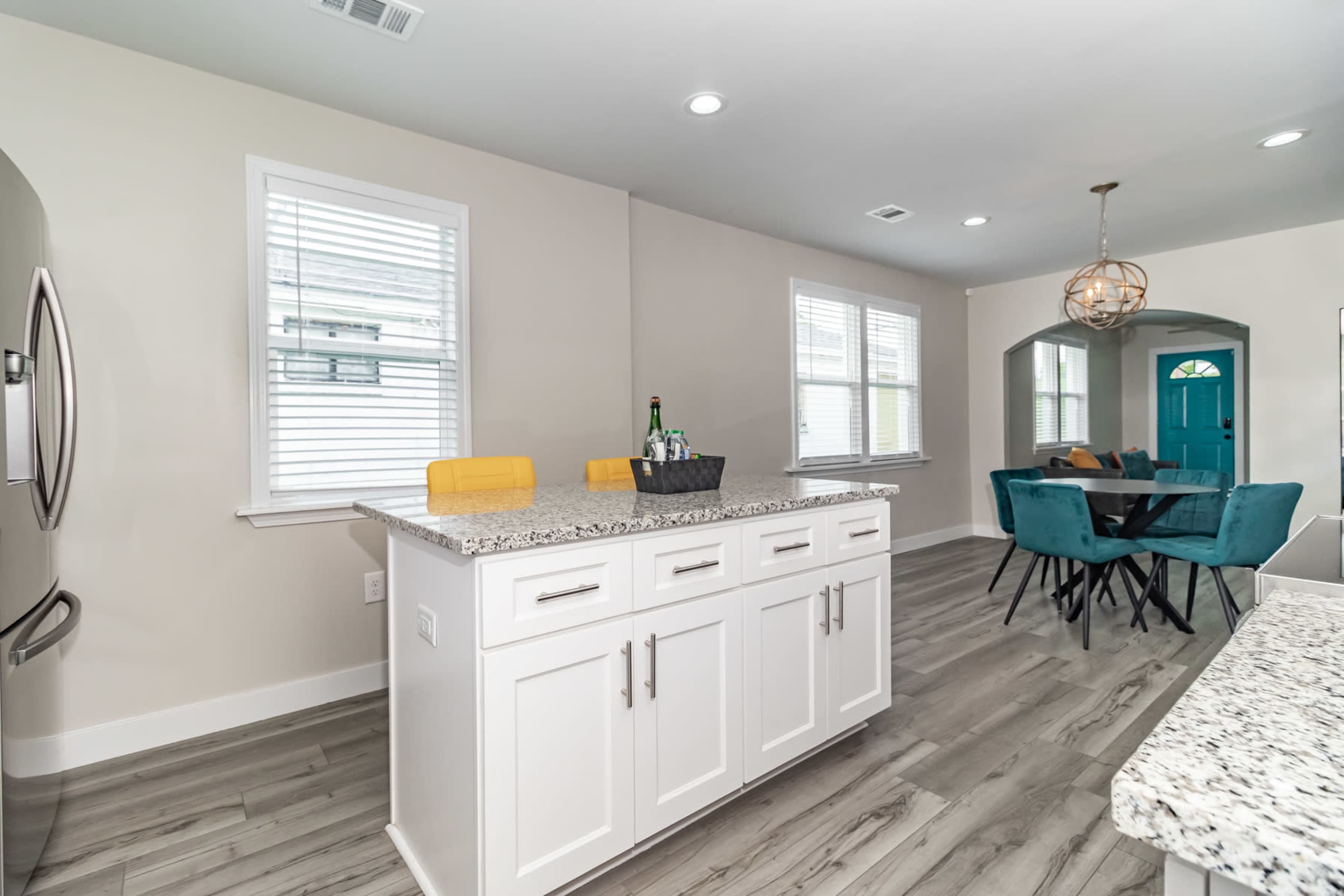 A modern kitchen features a central island with a granite countertop, stainless steel appliances, and a dining area with a blue door in the background.