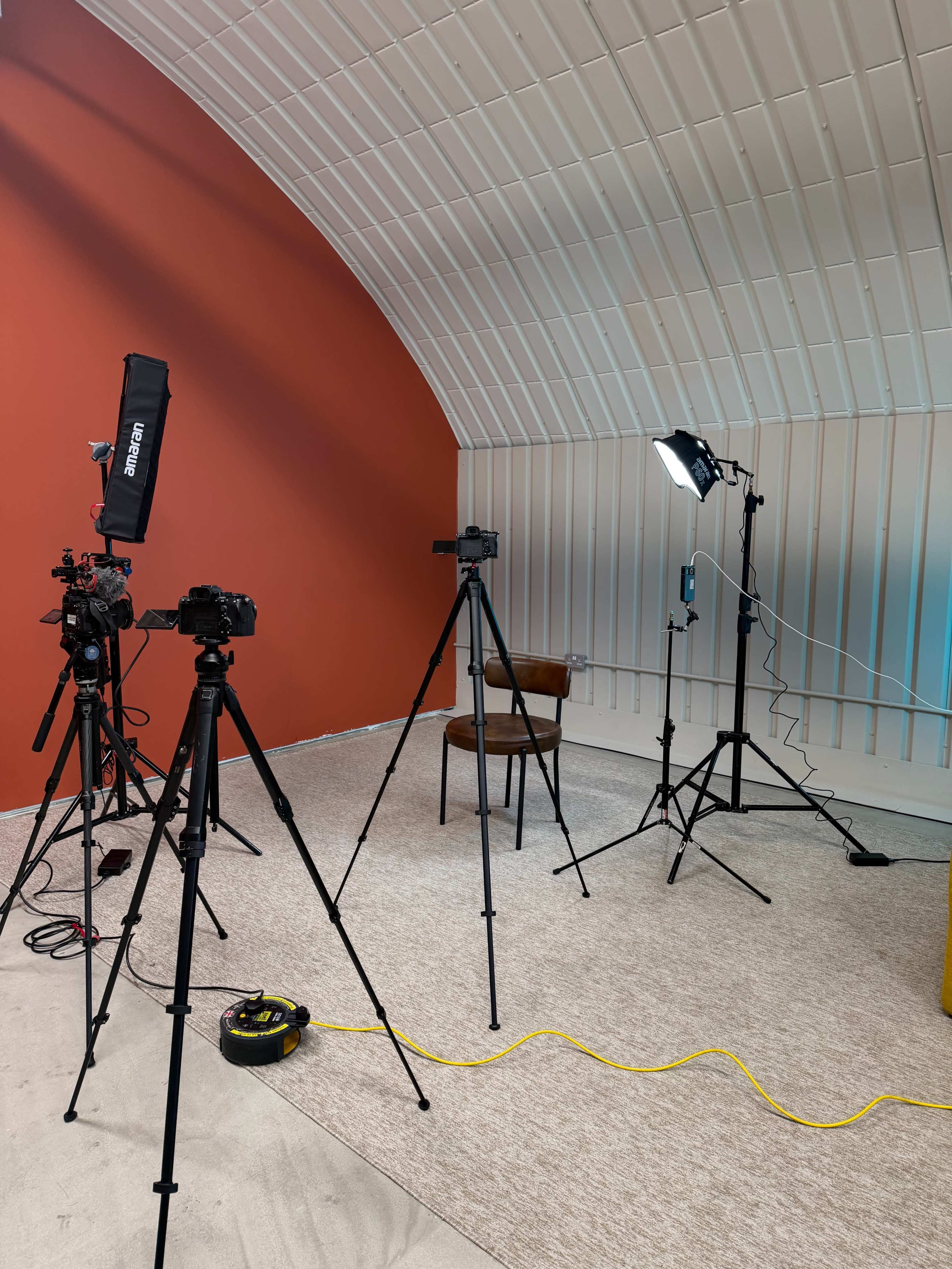 The image shows a plain room with a wooden chair set up in front of multiple camera tripods and lighting equipment against a vibrant red wall.