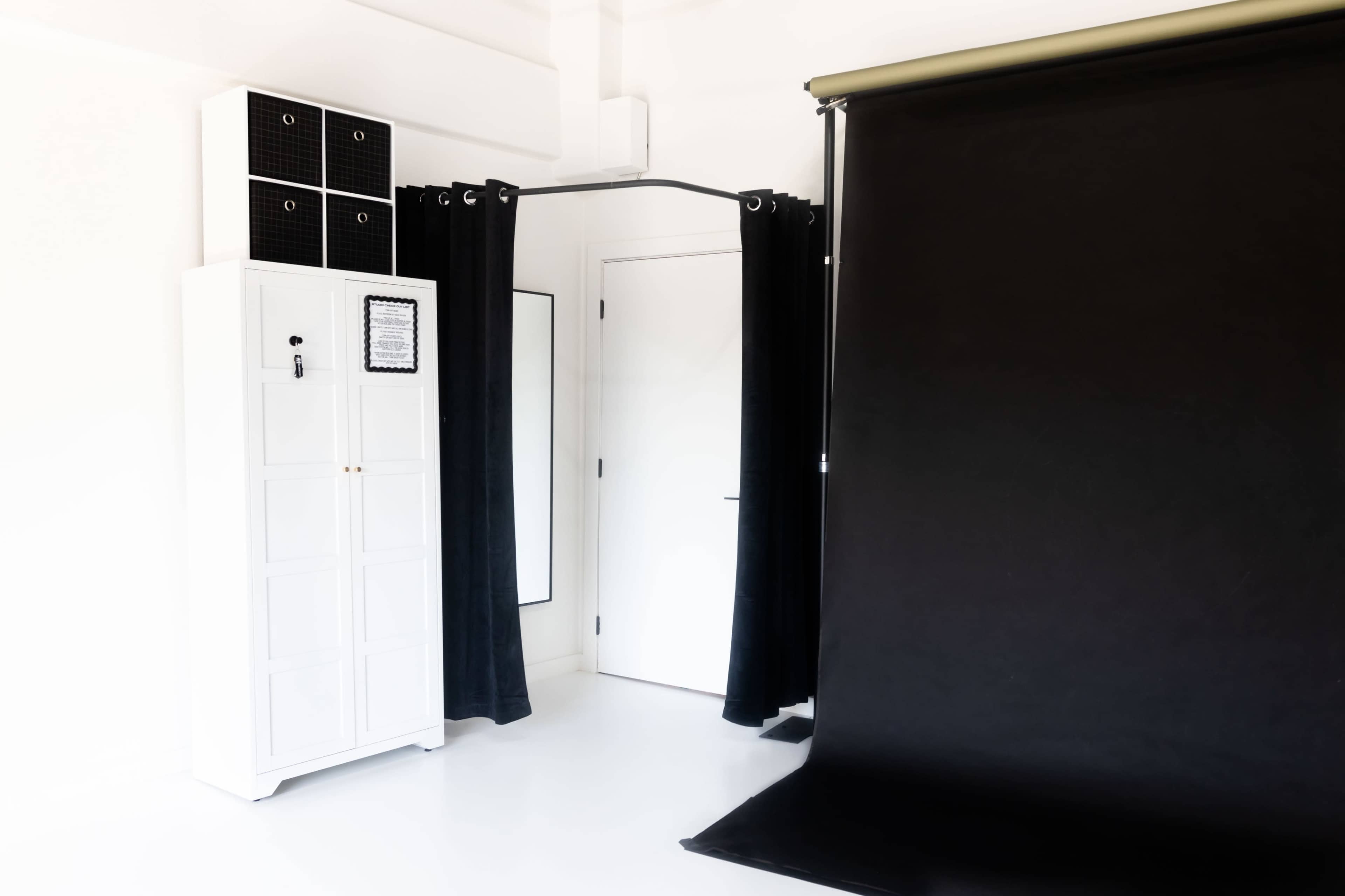 The image shows a minimalist studio space featuring a black backdrop, a white cabinet, and a mirror, with black curtains draping from a rail.