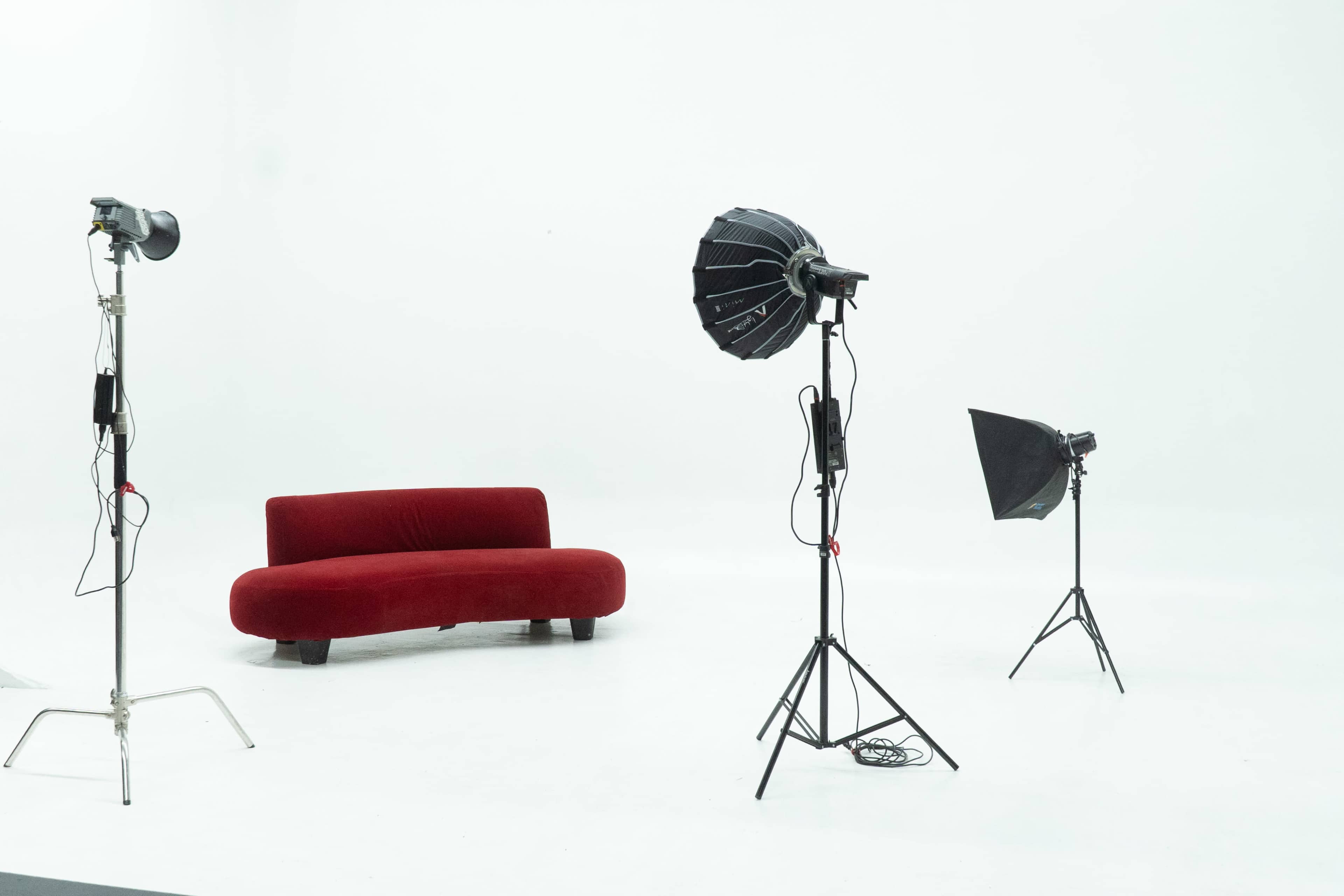 A red curved sofa is placed in a minimalist studio setting with three lighting stands positioned around it.