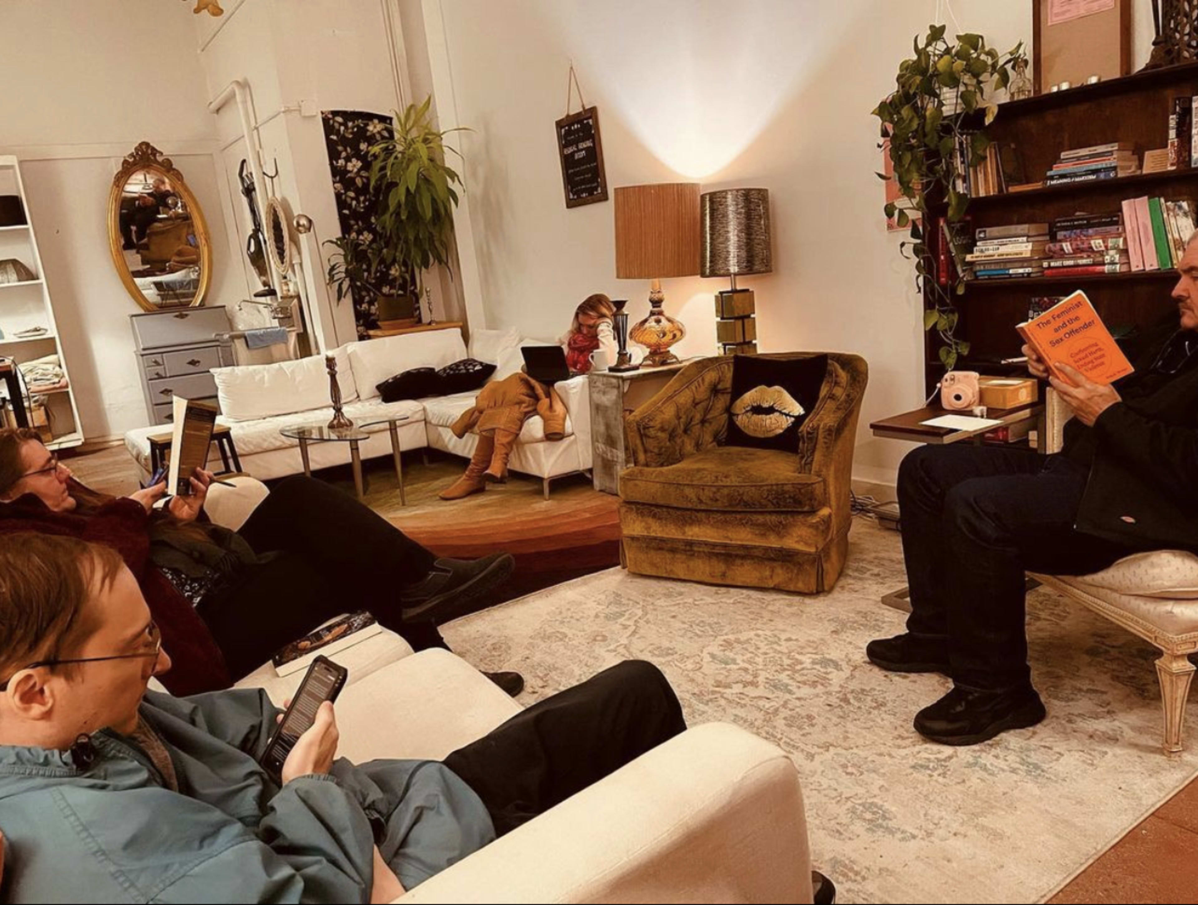 Four people are seated in a cozy, book-filled room, with one person reading a book while the others engage with their devices.