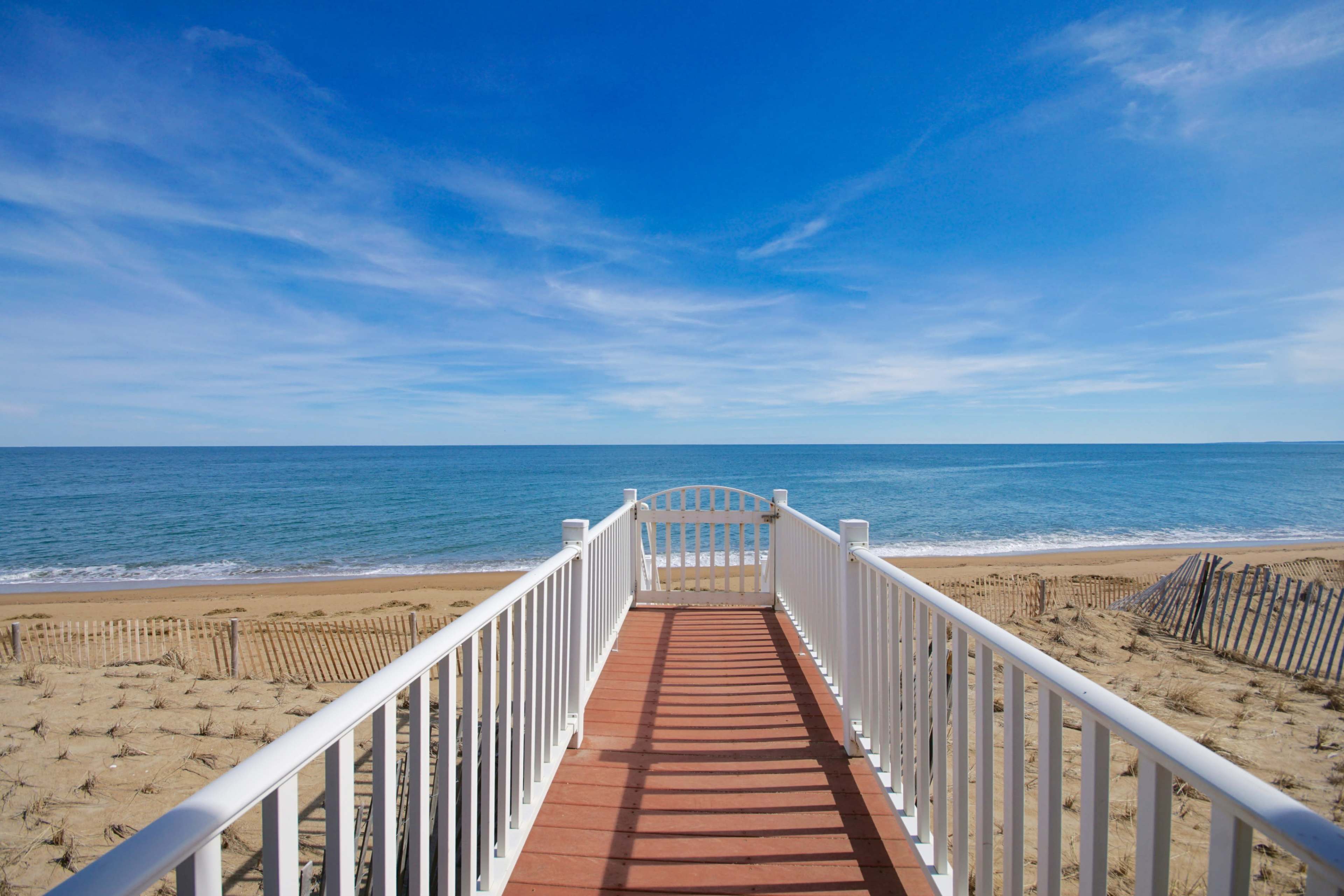 A white wooden walkway extends towards a sandy beach and calm ocean under a clear blue sky.