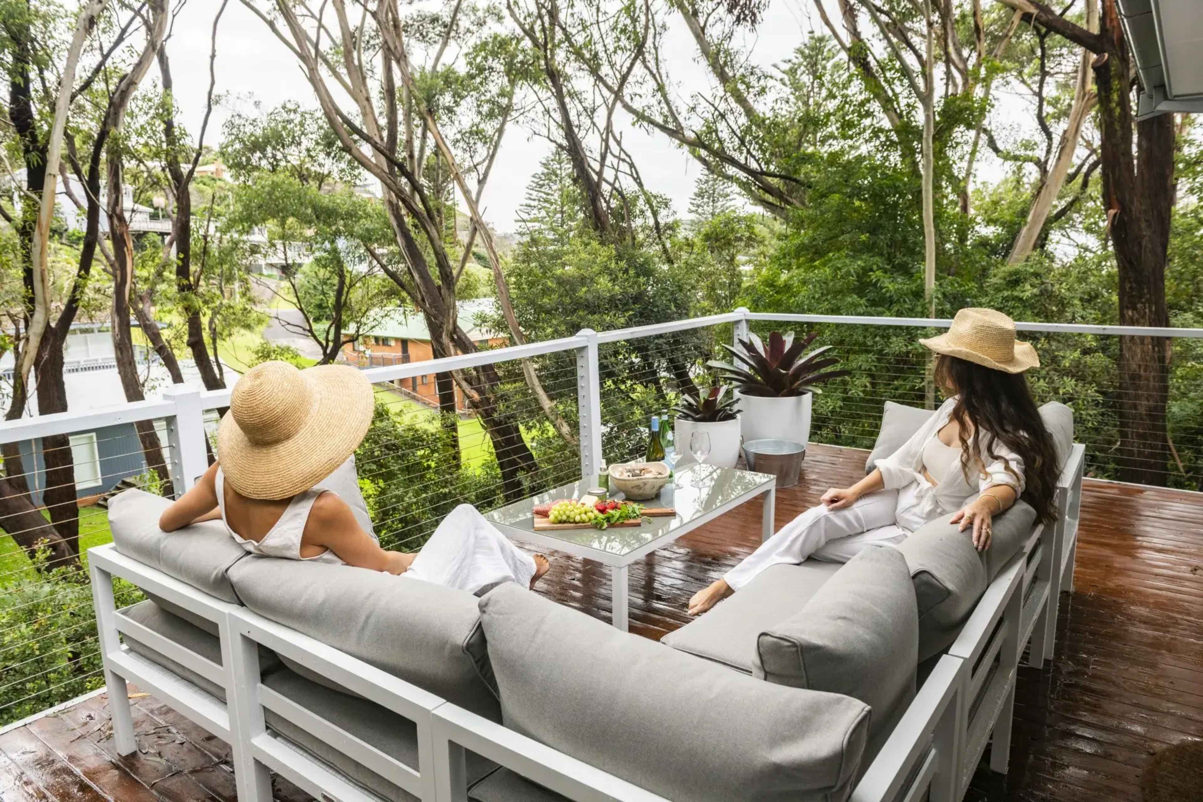 Two women wearing wide-brimmed hats sit on a furnished balcony surrounded by greenery, with a table displaying fruit and snacks.
