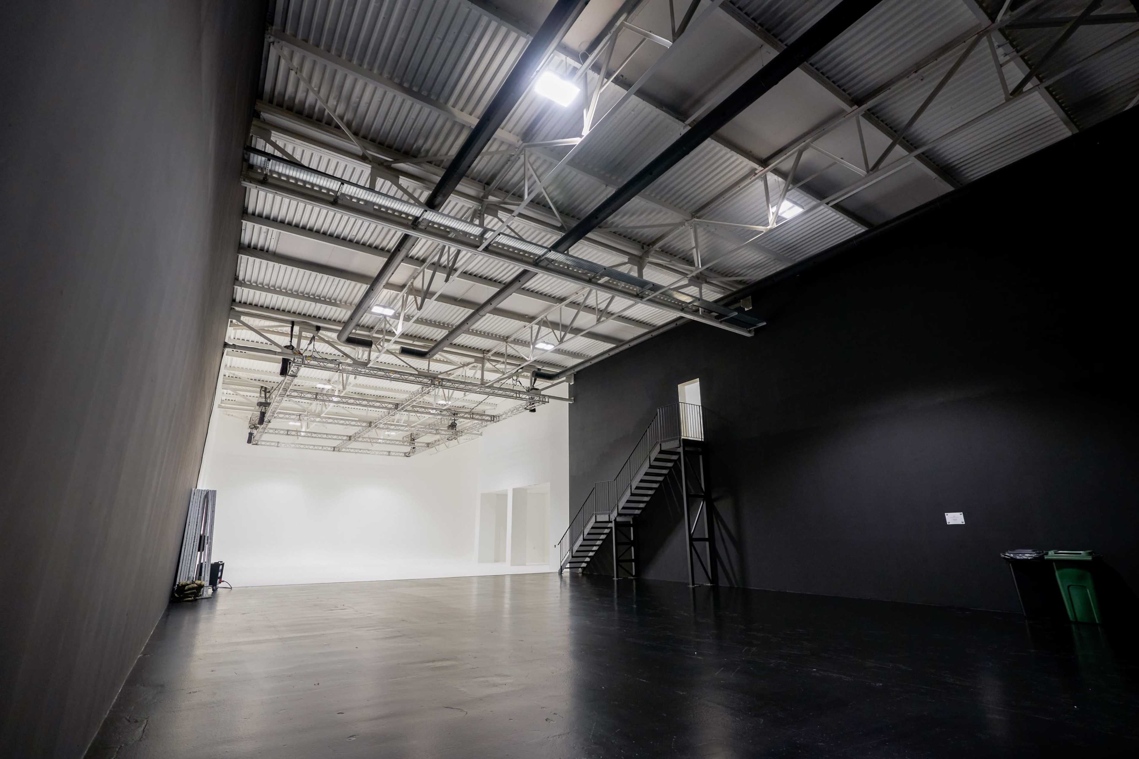 The image shows a spacious indoor area with a stark black wall, a white backdrop at the far end, and a staircase leading to a second level.