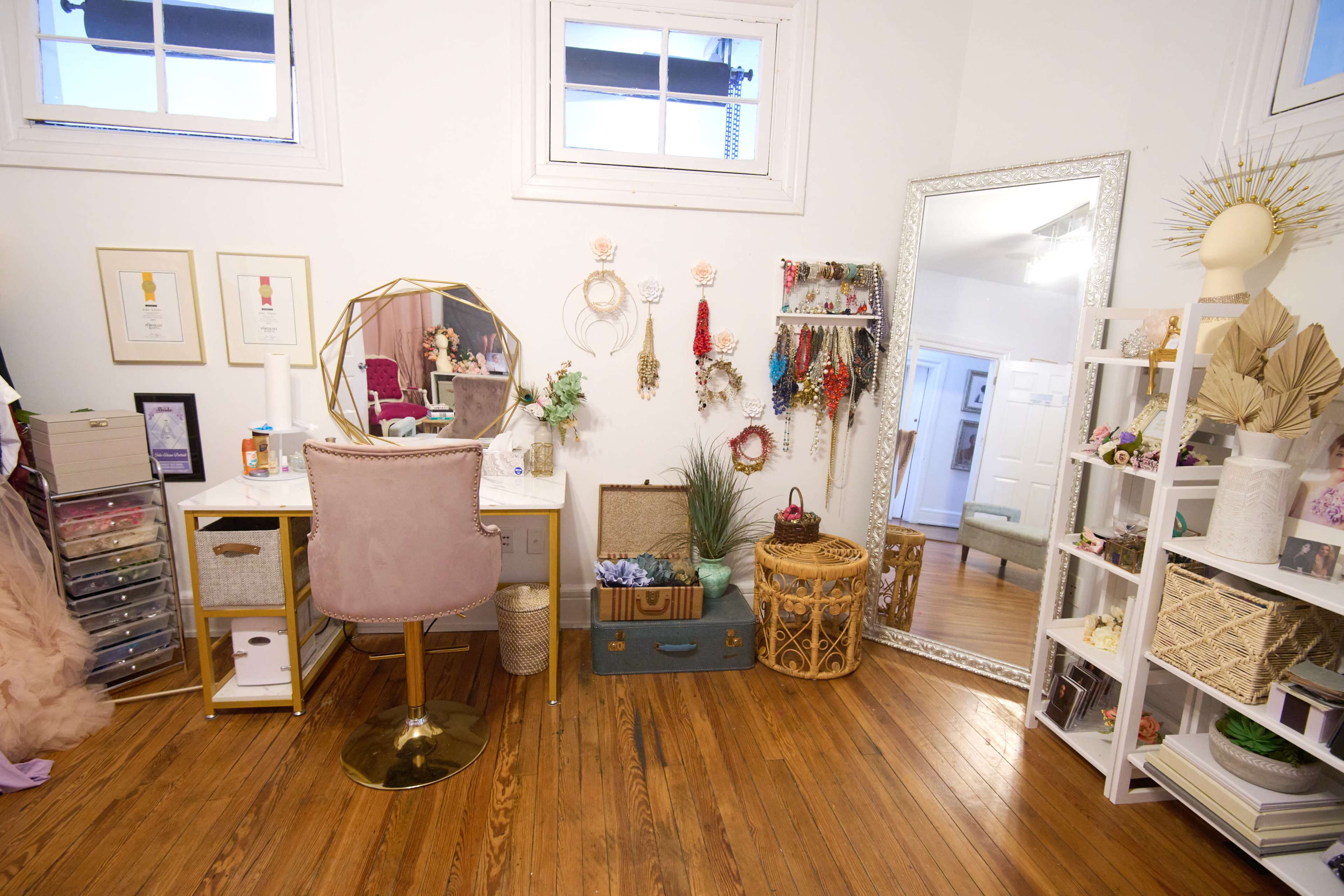The image shows a well-organized feminine workspace featuring a vanity desk with a chair, decorative wall hangings, a large mirror, and various storage solutions in a room with hardwood flooring.