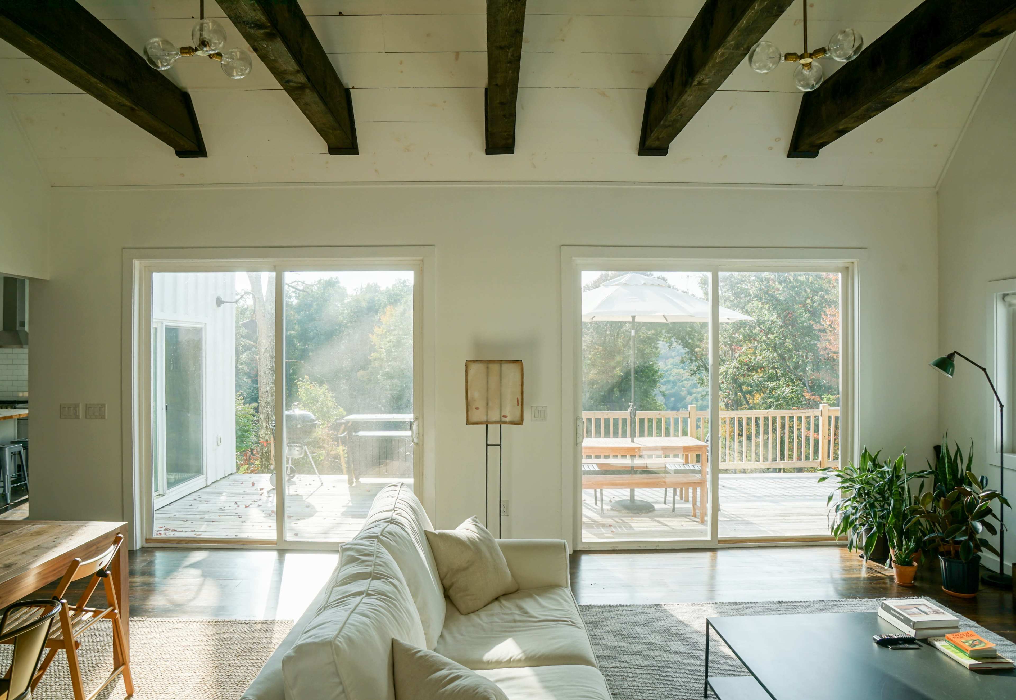 A bright living room with wooden beams features large glass doors leading to a deck with outdoor furniture.