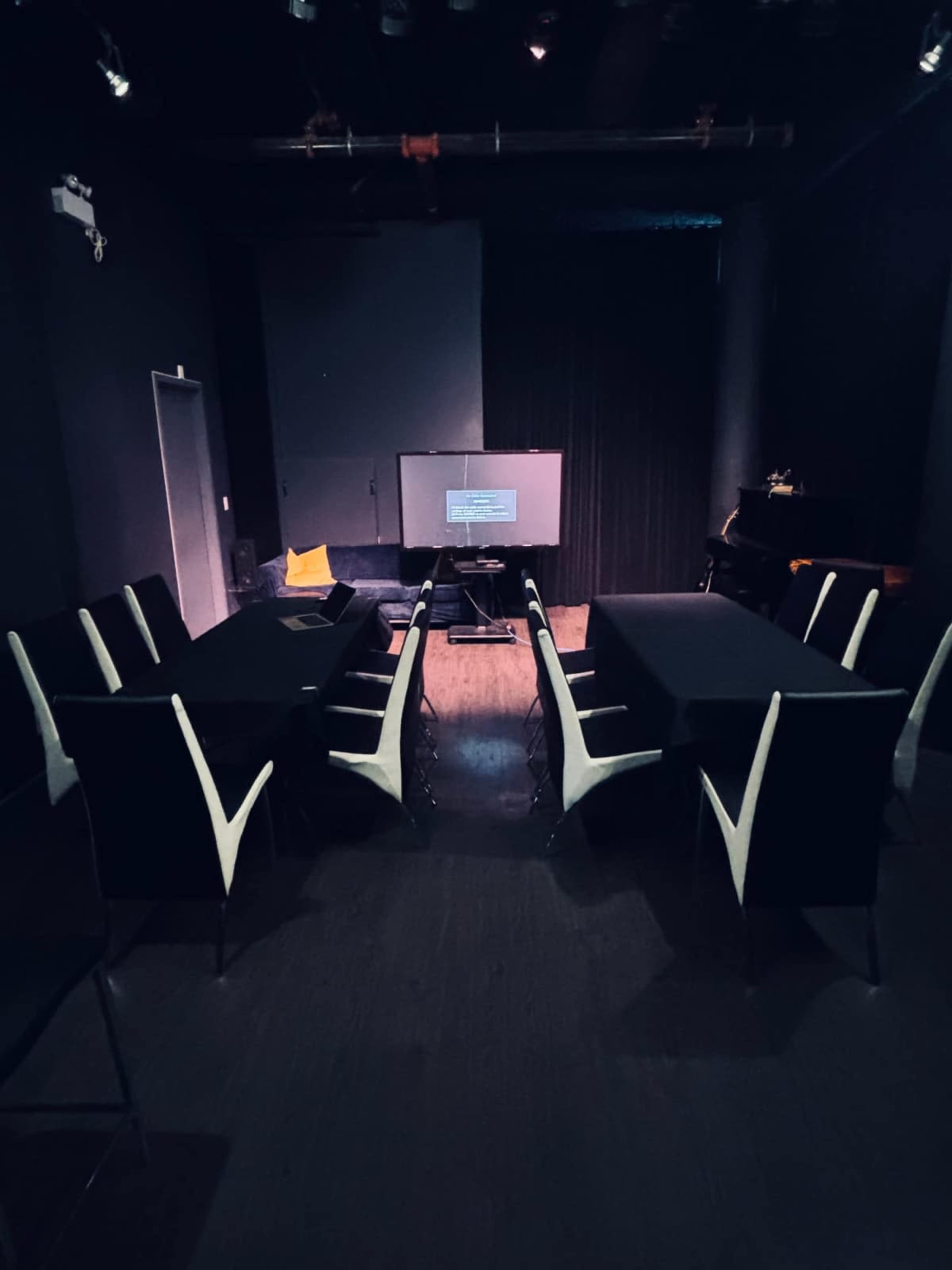 A dimly lit meeting room features rectangular tables arranged in a U-shape, with a projection screen at the front and a couch in the background.