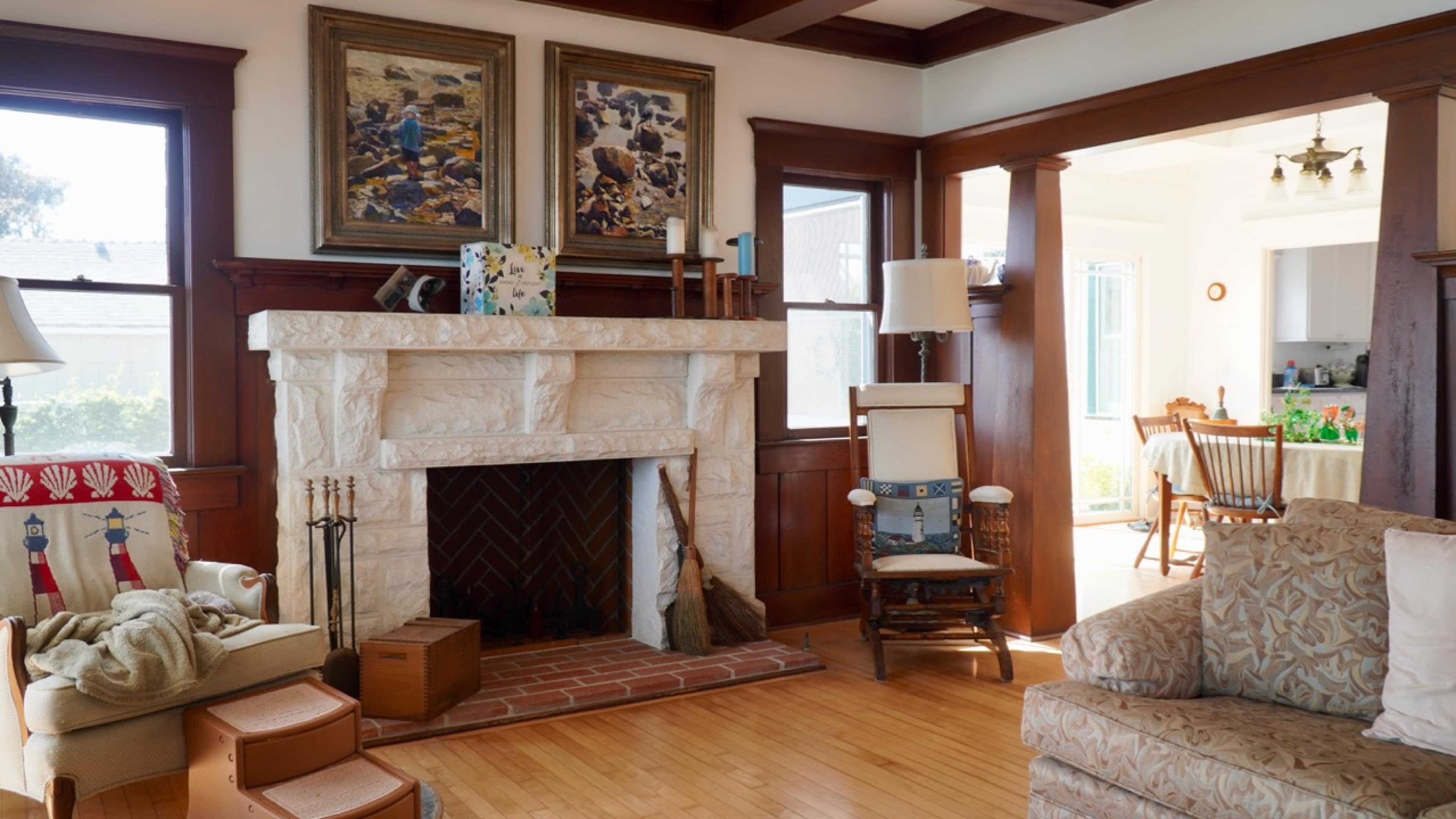 The image shows a cozy living room with a stone fireplace, wooden accents, and a view of a dining area in the background.