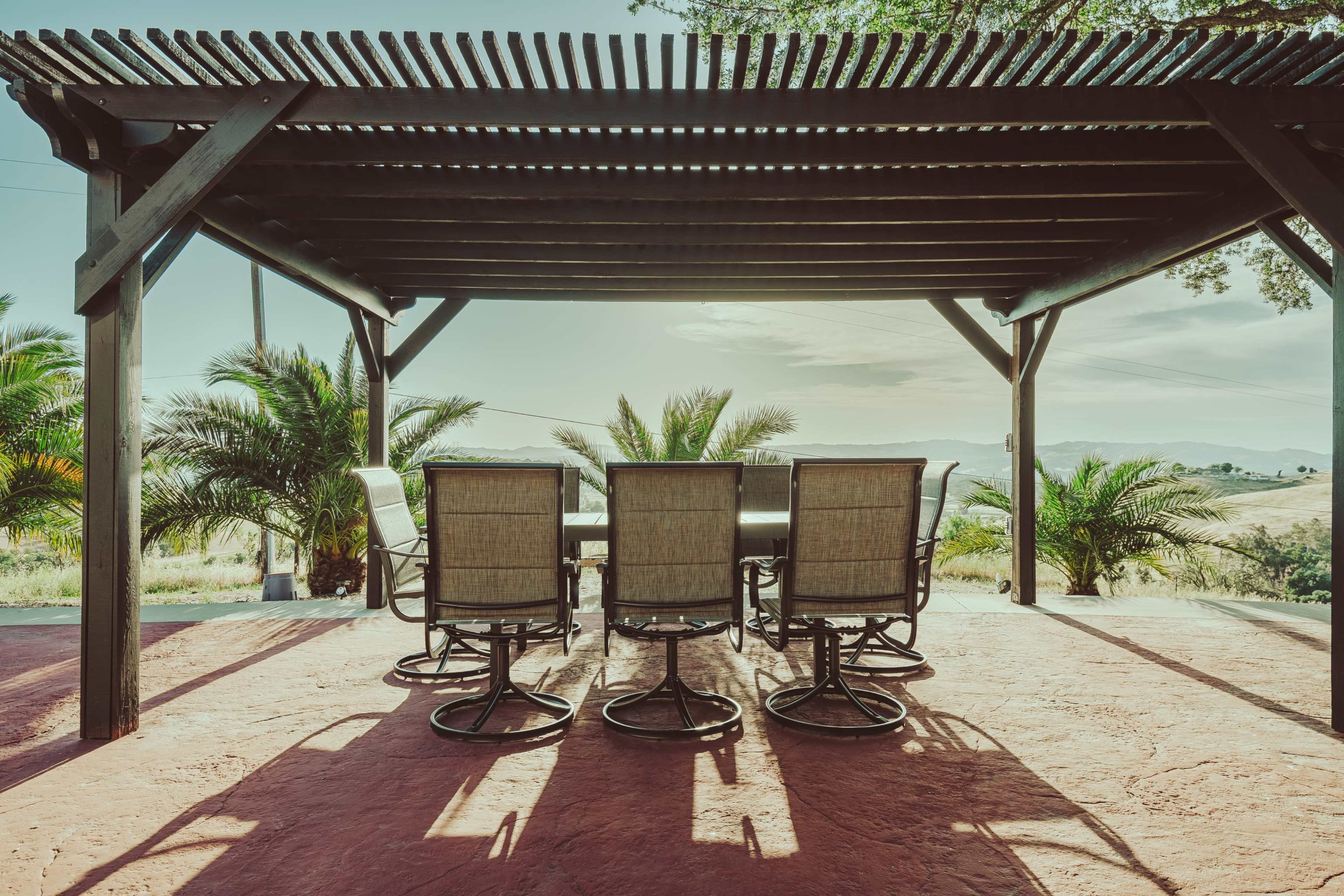 A patio with a table and chairs is positioned under a wooden pergola, overlooking a landscape with palm trees and rolling hills.