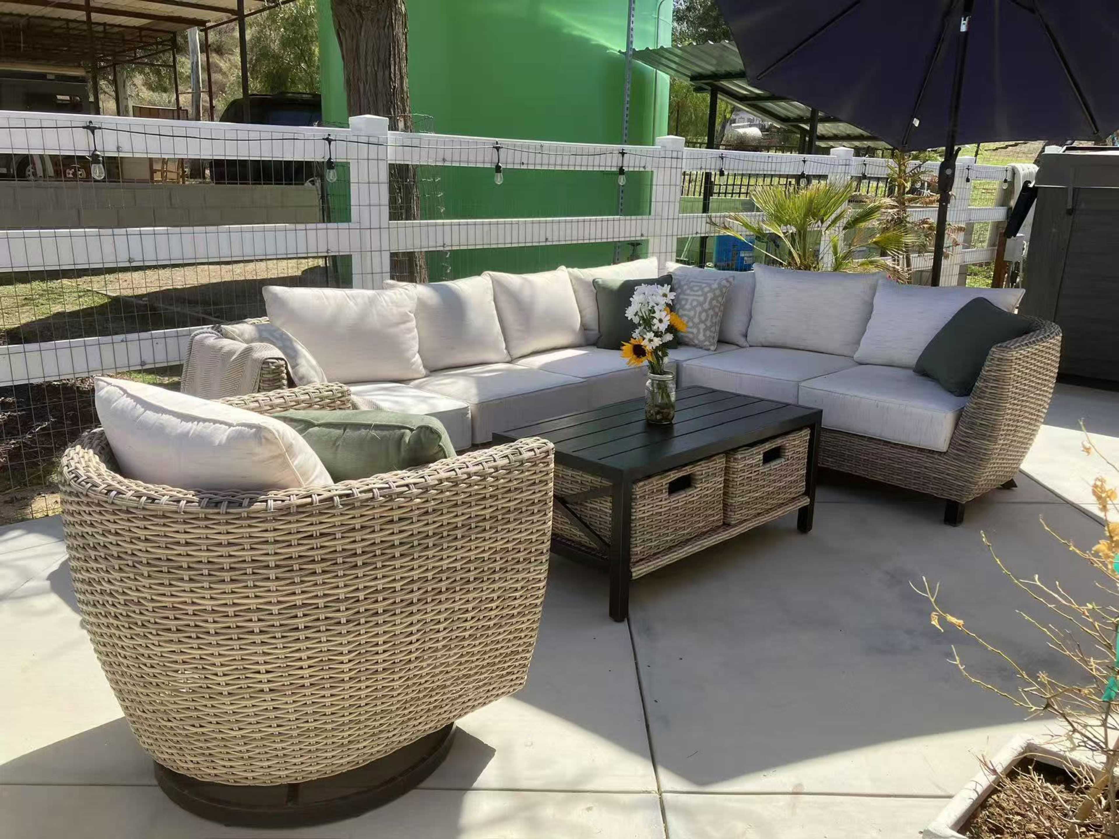The image shows an outdoor seating area featuring a sectional wicker sofa with cushions, accompanied by a round wicker chair and a black coffee table, all set on a concrete surface.