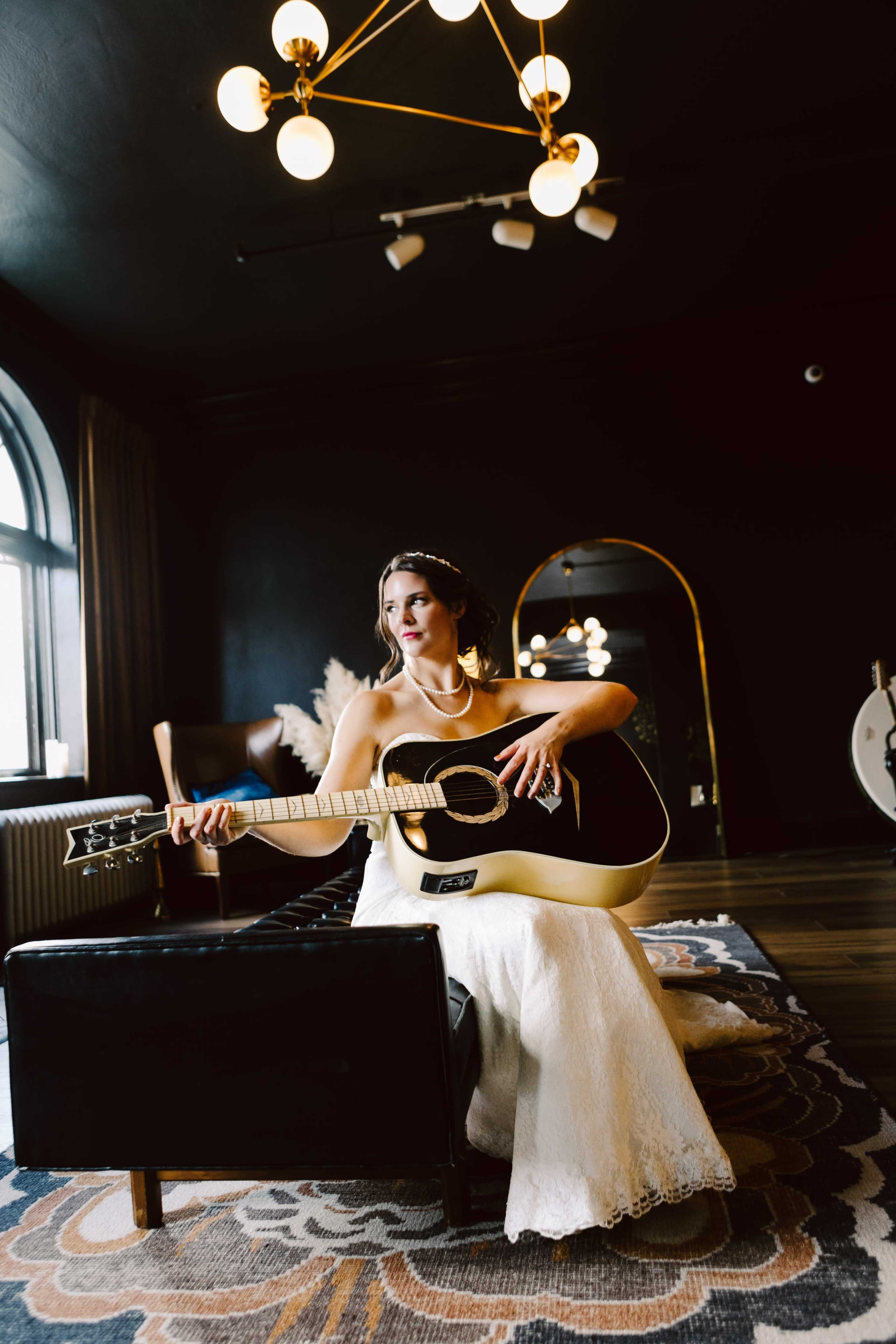 A bride in a lace wedding gown sits on a black bench, holding an acoustic guitar in a dark, modern room with stylish light fixtures.