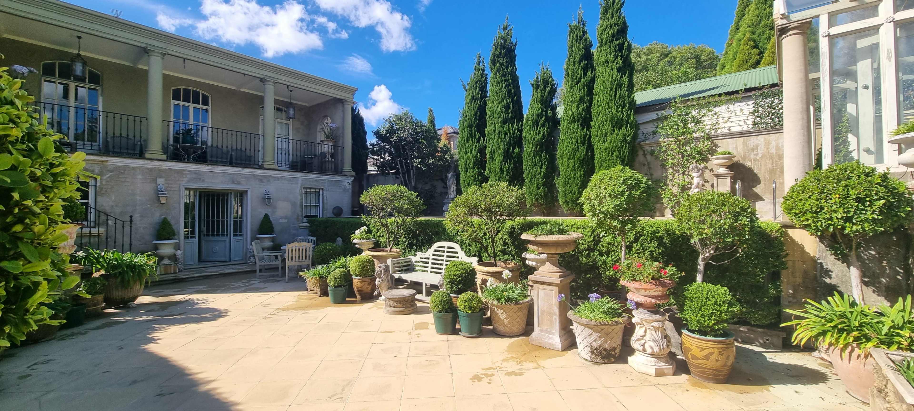 A stone courtyard features potted plants and small trees surrounding a set of seating areas, with a two-story building and blue sky in the background.