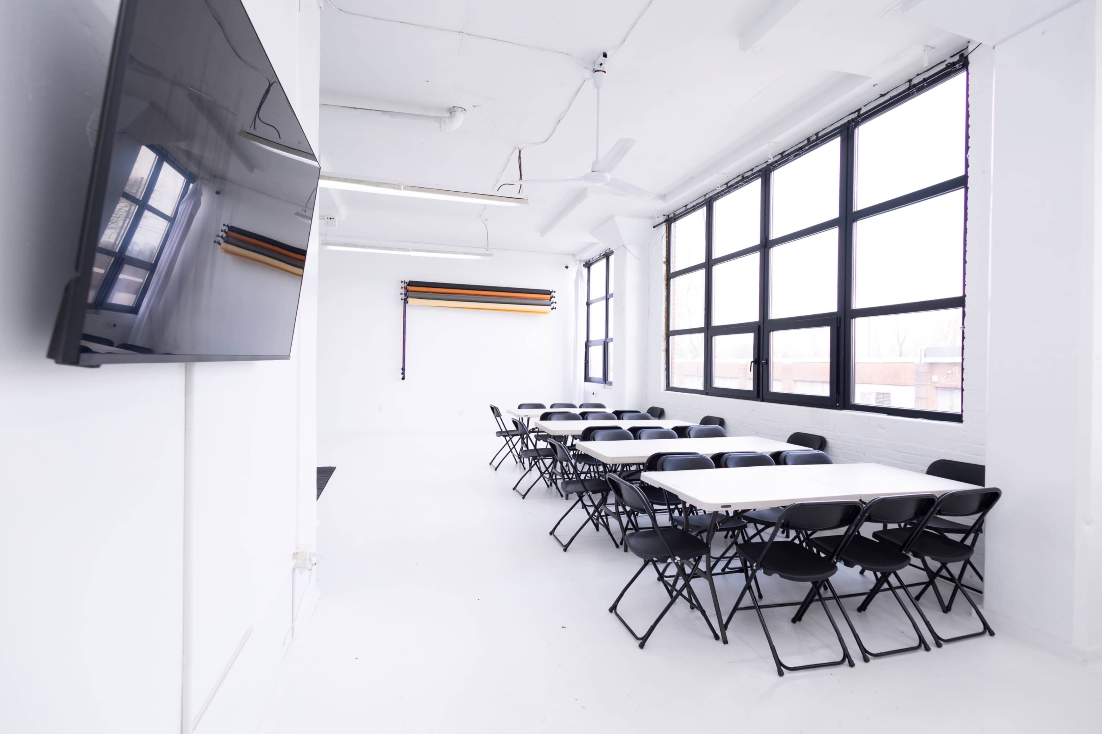 The image shows a bright, modern meeting room with multiple tables arranged in a long configuration and black chairs.