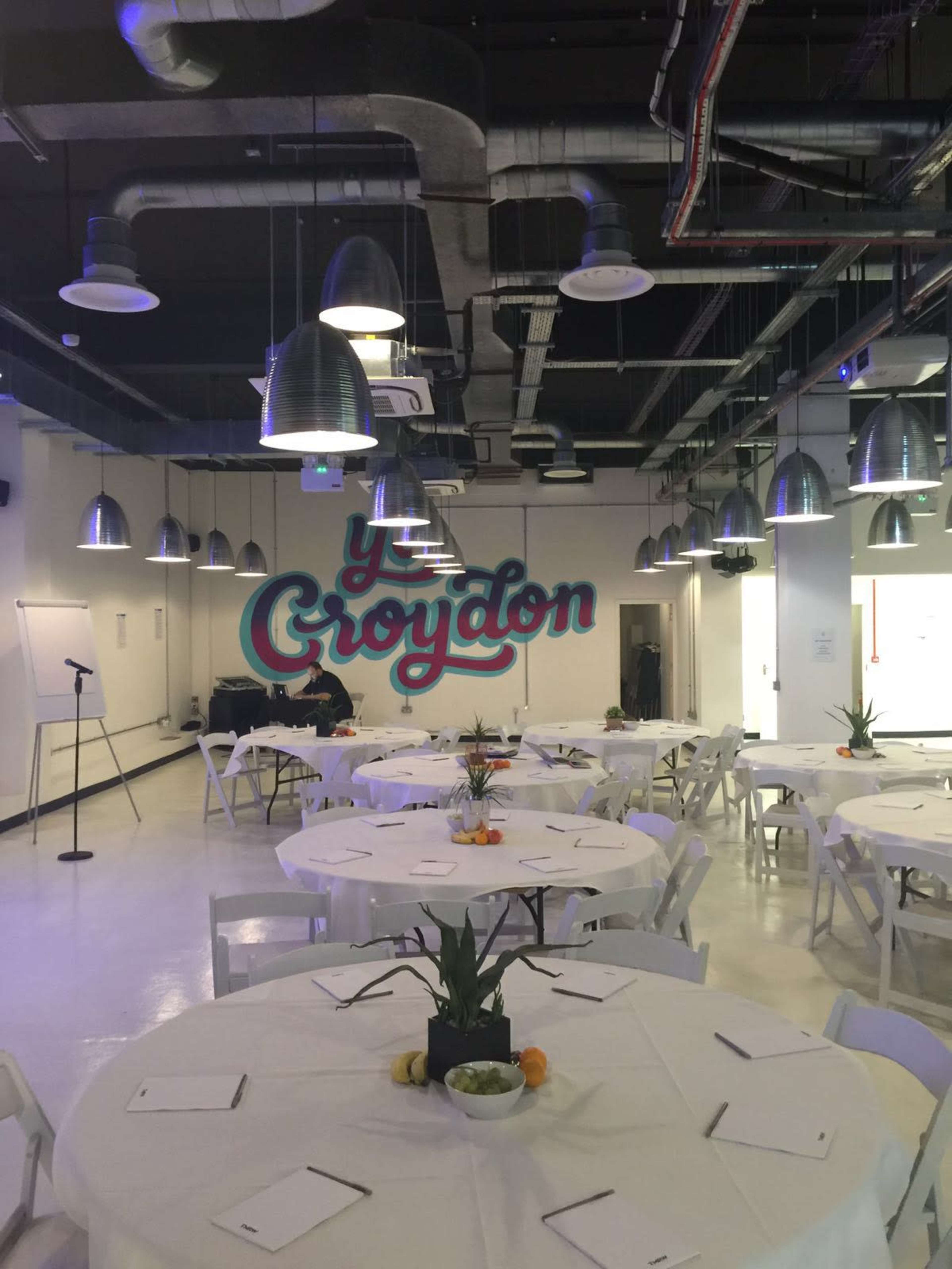 A modern event space with round tables set for a gathering, featuring a mural that says "y Croydon" on the wall and industrial-style hanging lights above.
