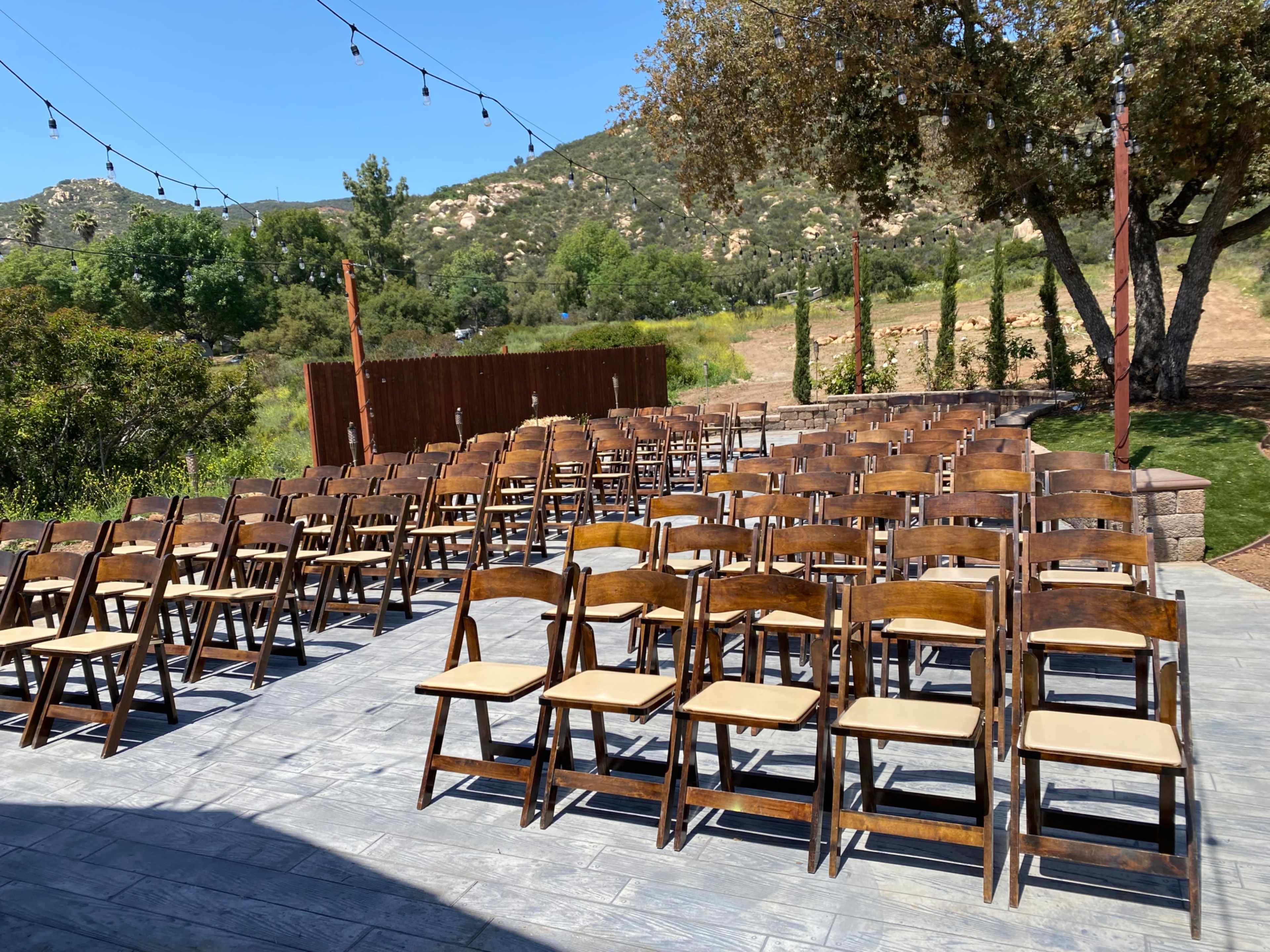 A set of wooden chairs arranged in rows on a patio, with a backdrop of trees and rolling hills under a clear blue sky.