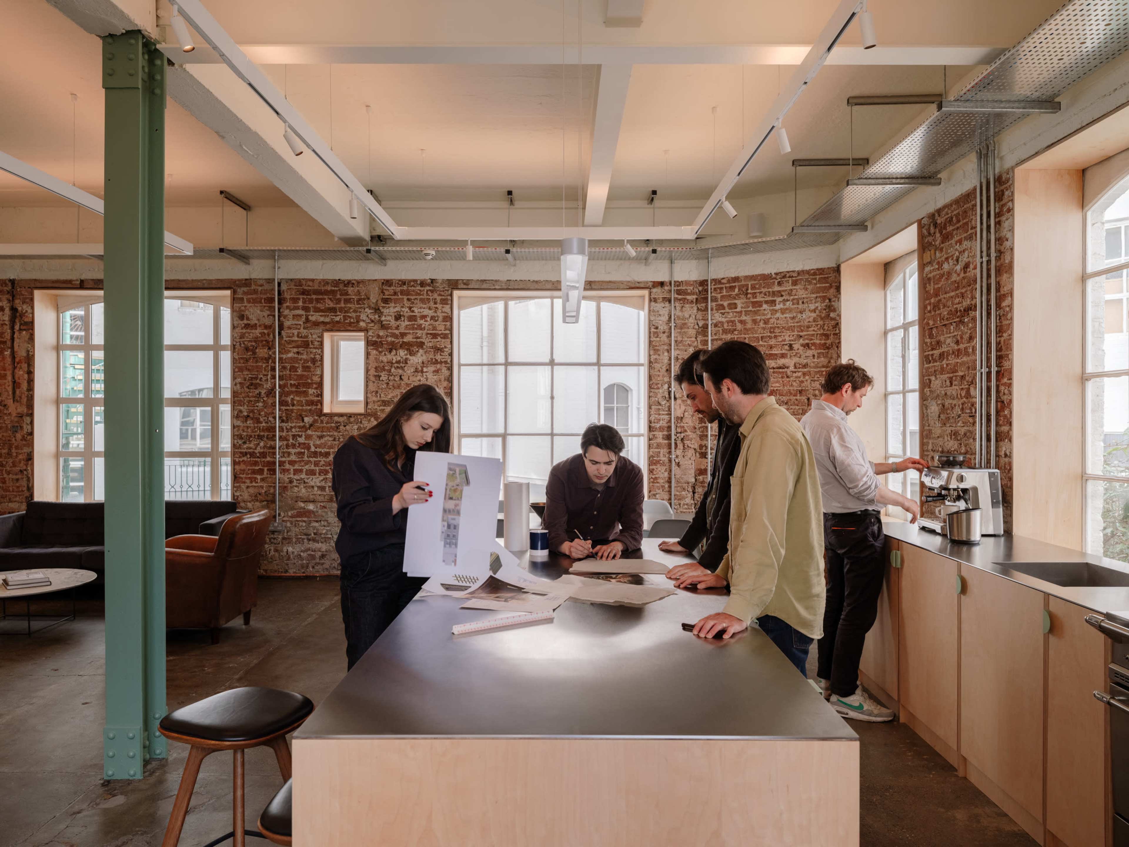A group of five people collaborate around a table in a modern office space with exposed brick walls and large windows.