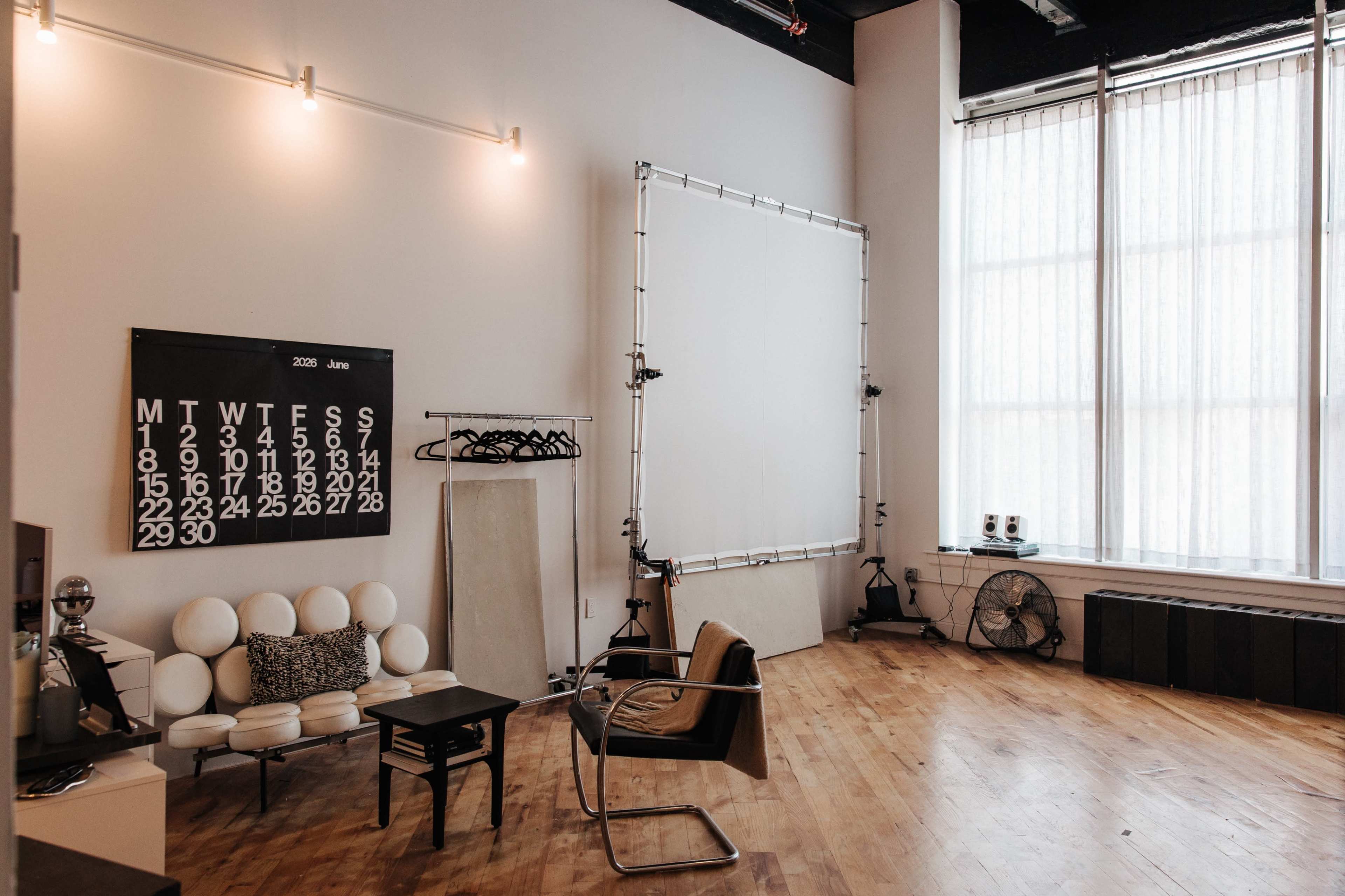 The image shows a spacious, minimalistic studio with a large window, a calendar on the wall, and furniture including a white couch and a brown armchair.