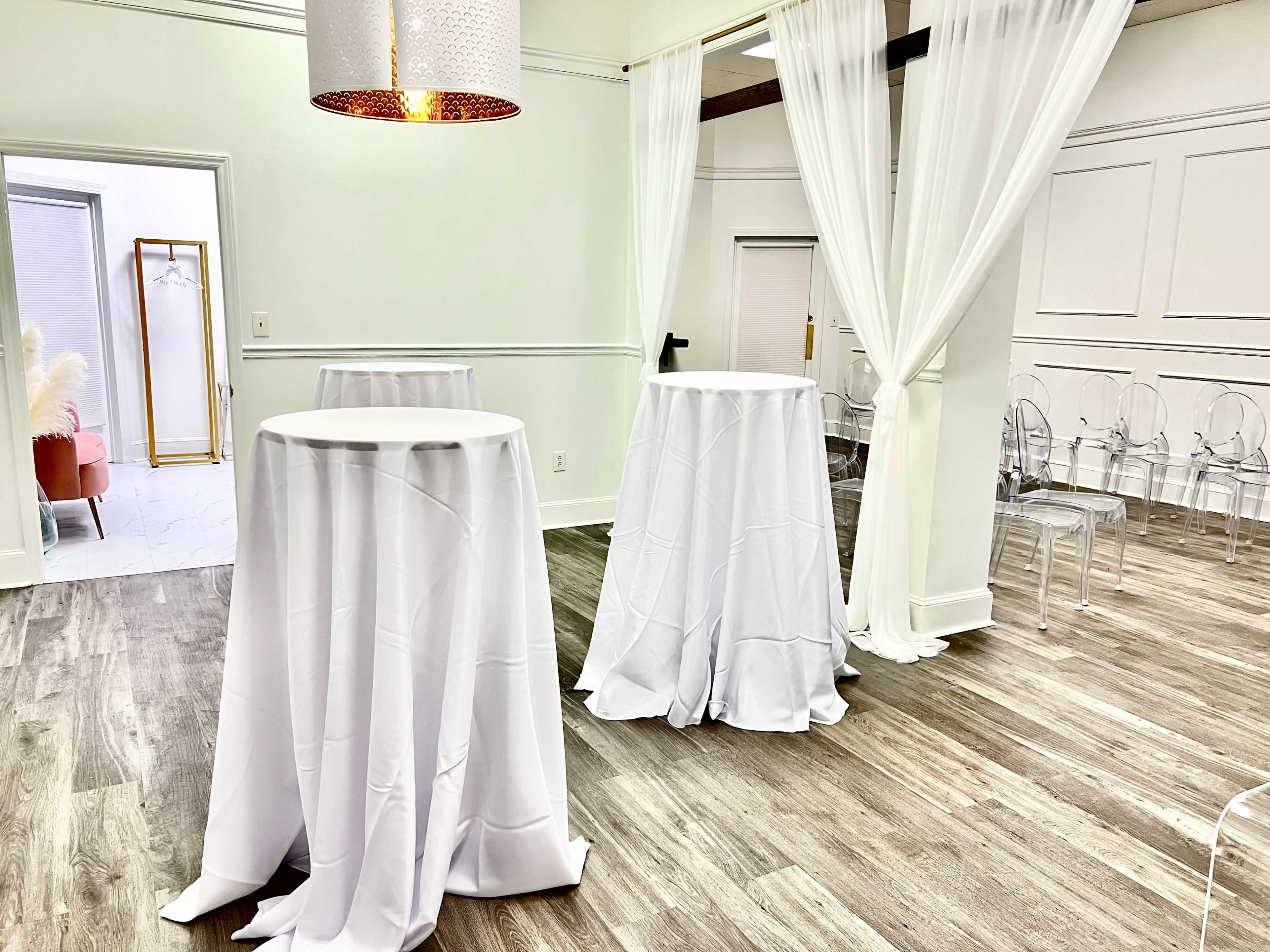 A bright event space features several tall, white draped tables and clear chairs arranged against a light-colored backdrop.