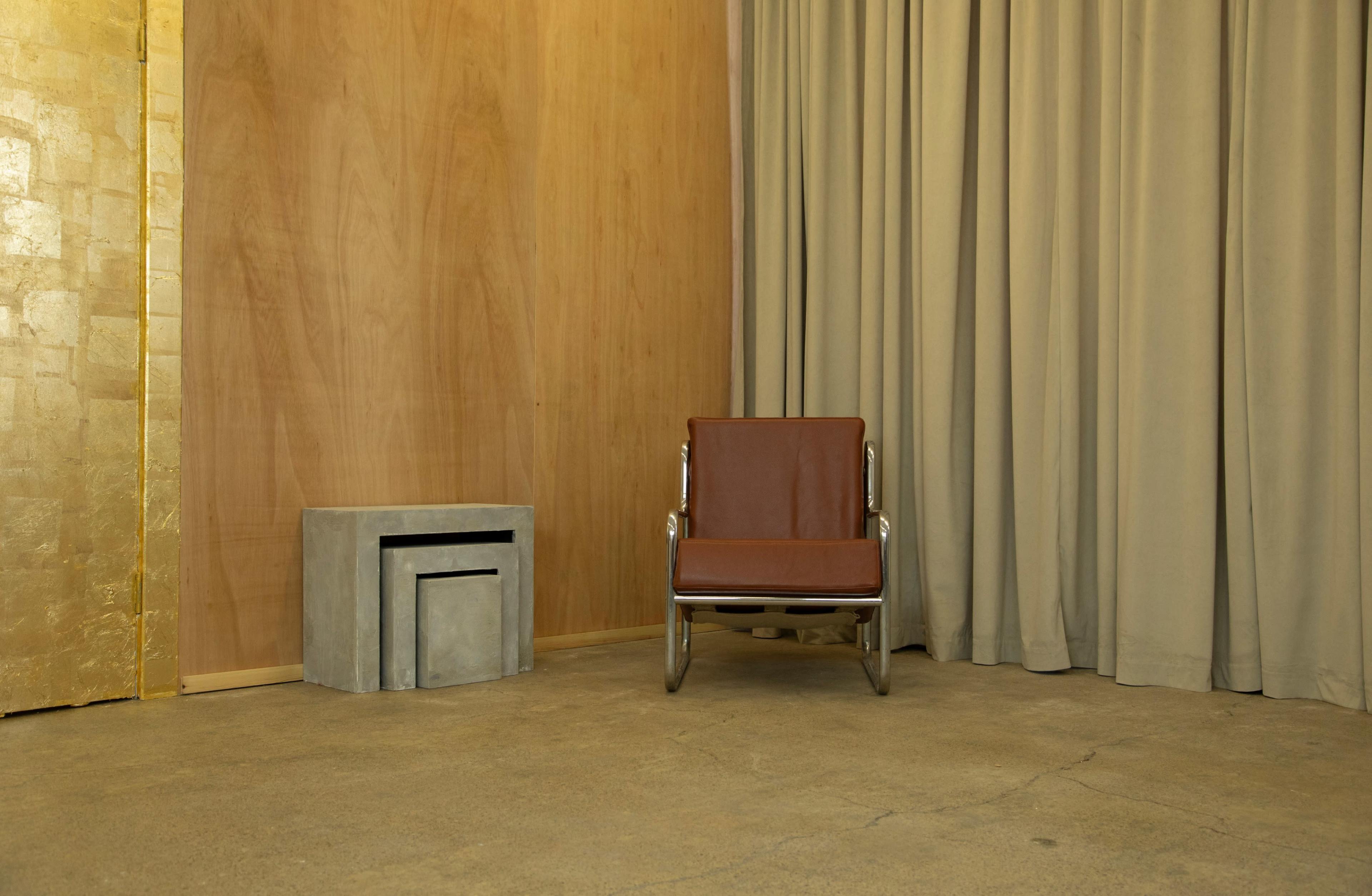 A minimalist room with a brown chair next to a concrete side table and a wall covered in light brown wooden panels, with a draped curtain in the background.