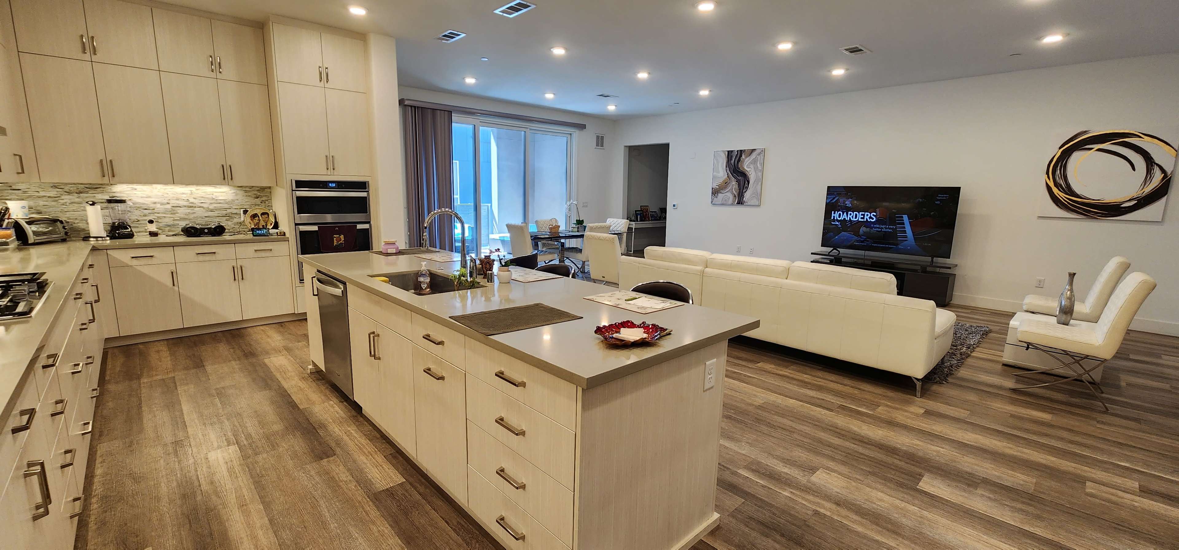 A modern kitchen and living area feature light-colored cabinetry, a central island, and a television mounted on the wall in an open-concept space.