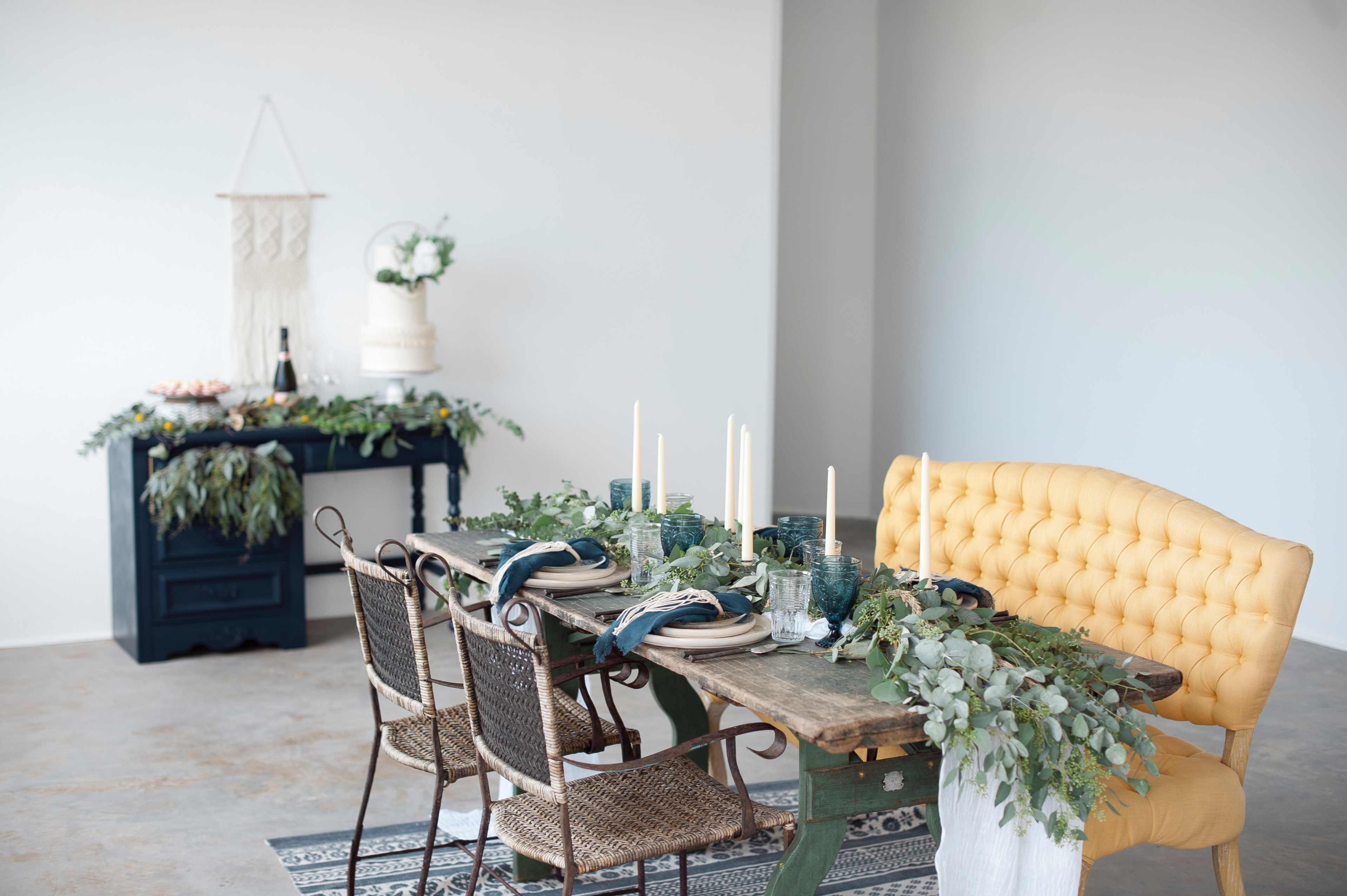 A rustic dining table is set with green foliage, candles, and tableware, alongside a yellow tufted loveseat in a well-lit room.