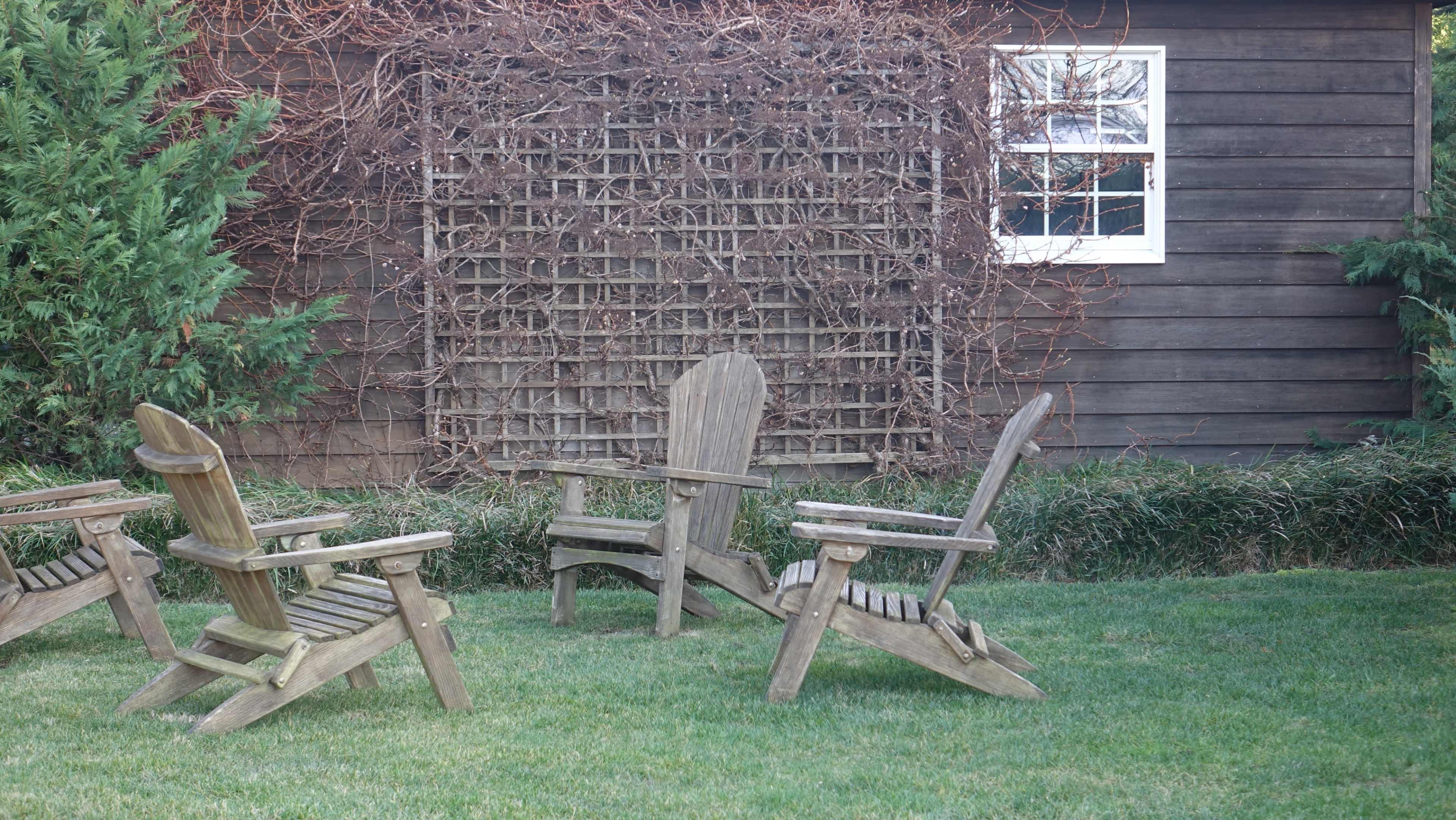 A set of wooden Adirondack chairs is arranged on a grassy lawn next to a wooden wall covered with a trellis of vines.
