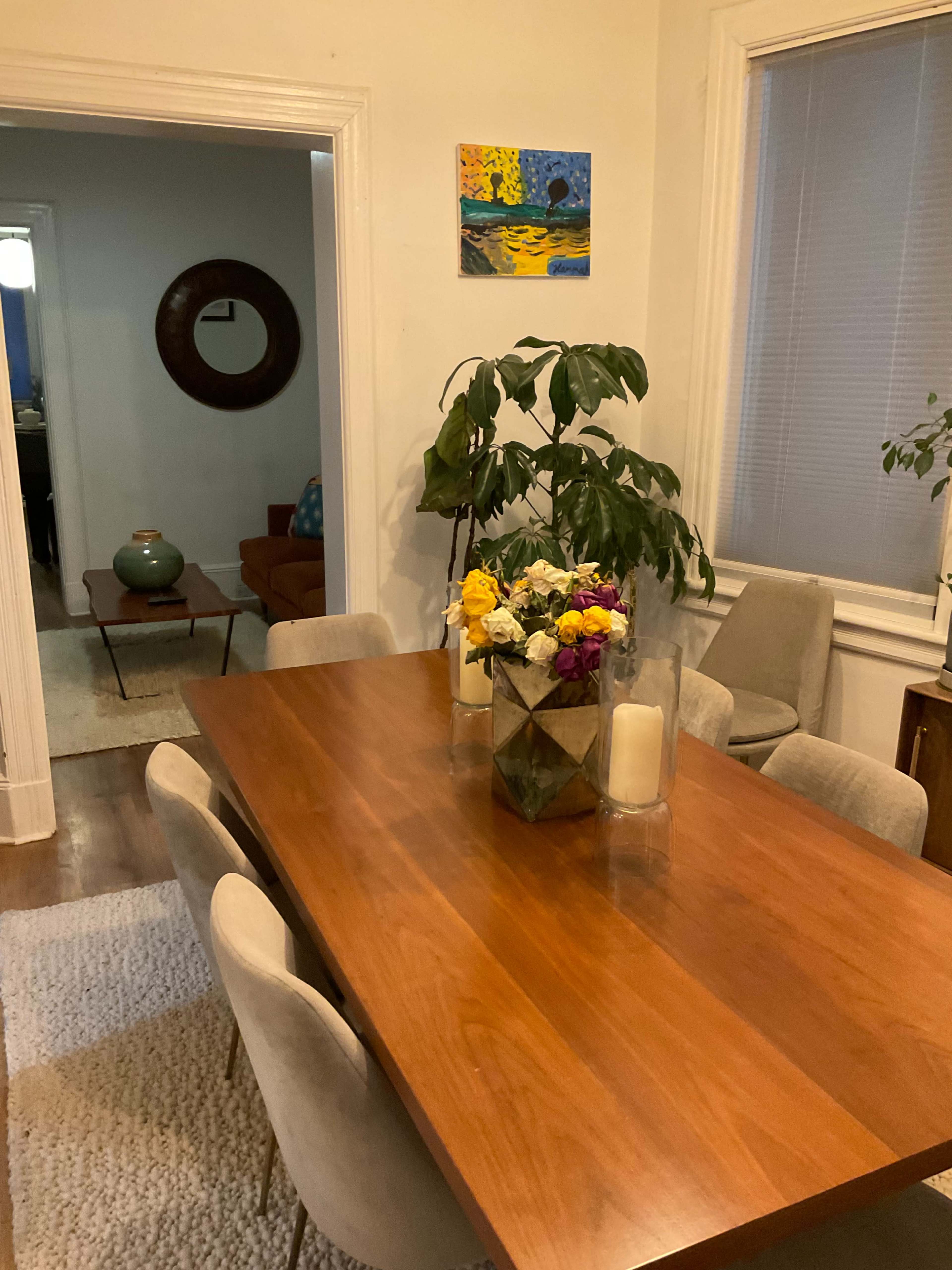 The image shows a dining area with a wooden table, surrounded by upholstered chairs, a vase of flowers in the center, and a doorway leading to another room in the background.