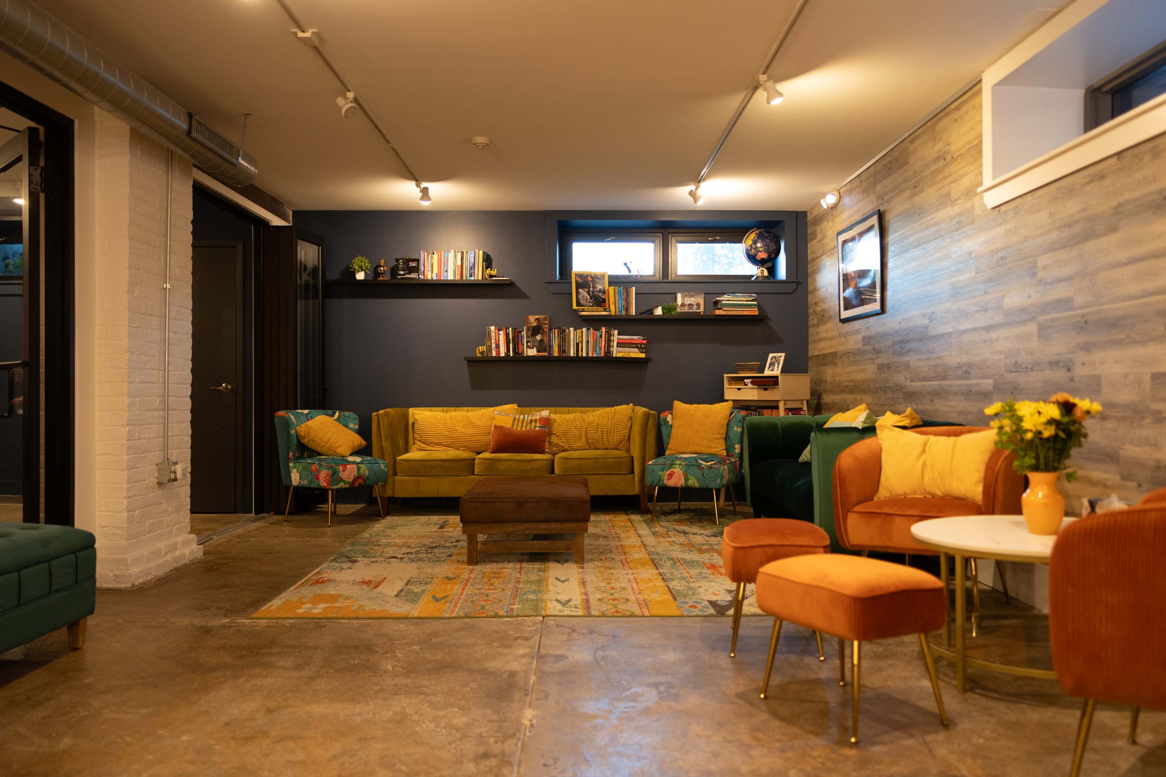 The image shows a cozy common area featuring various seating arrangements, a bookshelf, and decorative elements against a rustic wall.