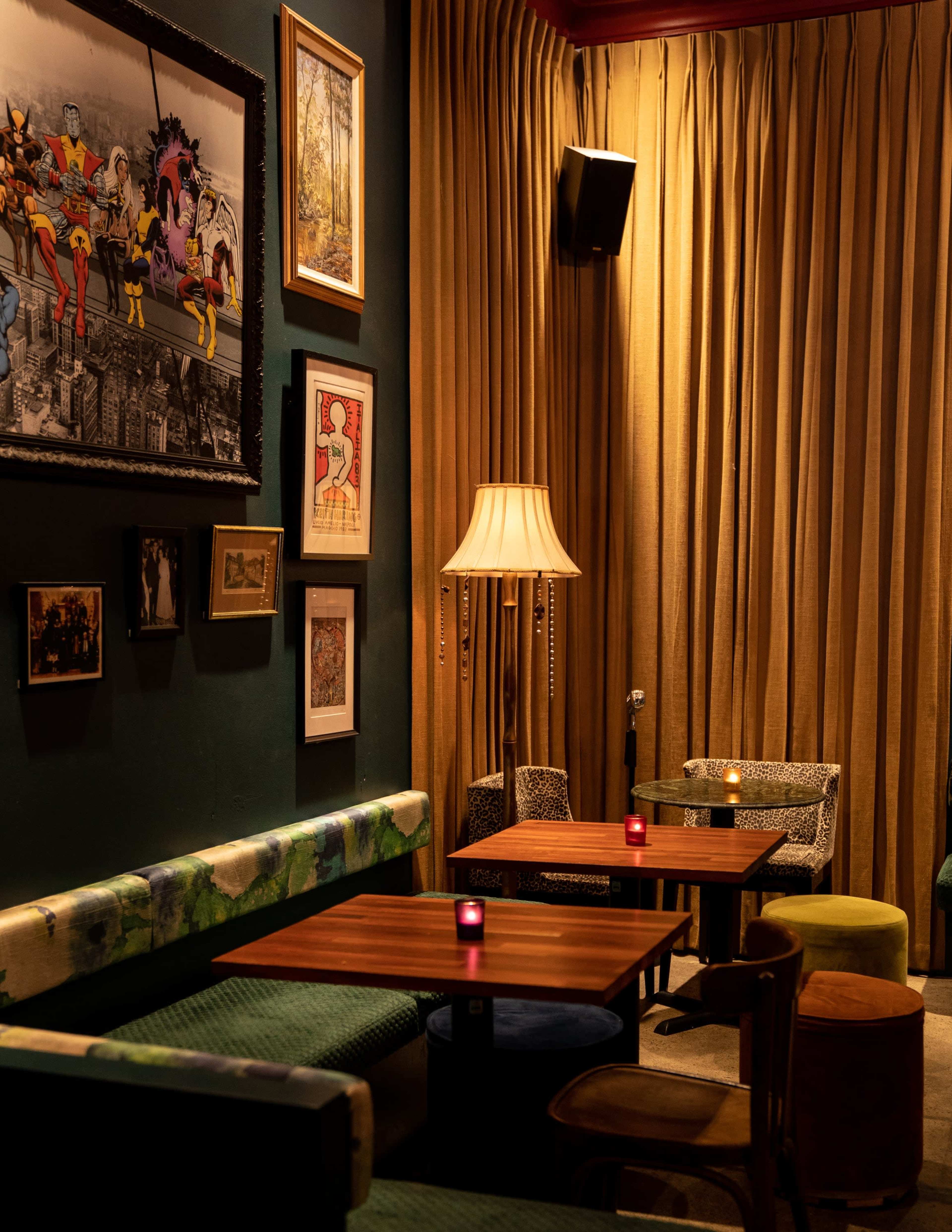 The image shows a cozy lounge area featuring patterned seating, a wooden table, artwork on the walls, and soft ambient lighting from a lamp.