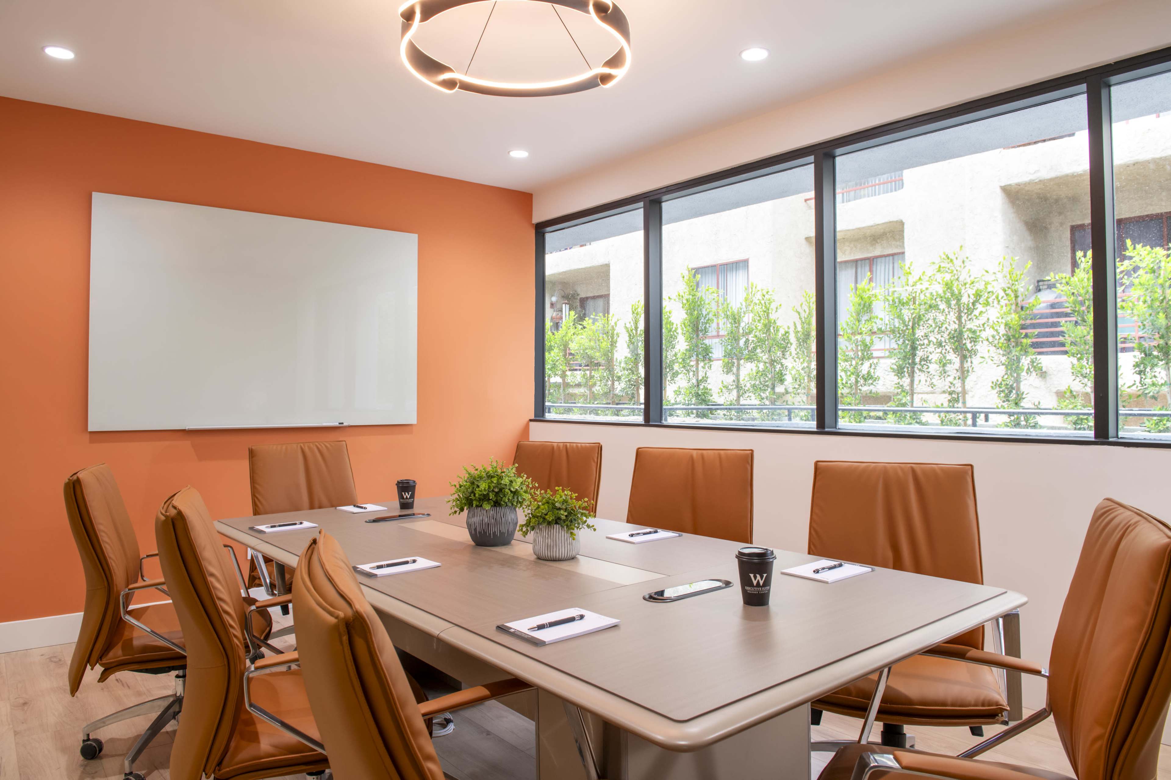 A modern conference room features a large table surrounded by orange leather chairs, with a whiteboard and large windows overlooking greenery.