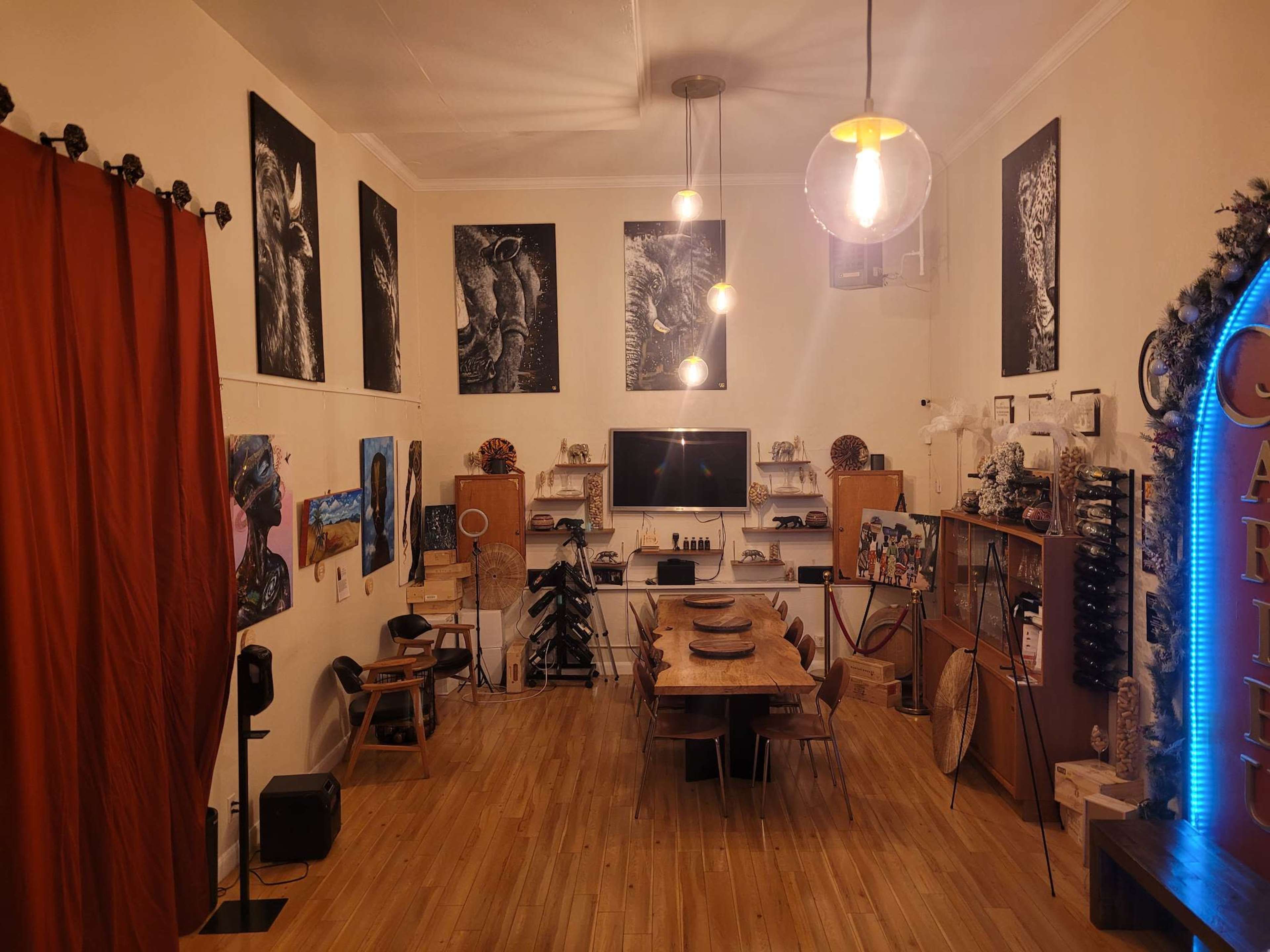 The image shows a spacious art studio featuring wooden furniture, several artworks on the walls, and a central dining table surrounded by chairs.