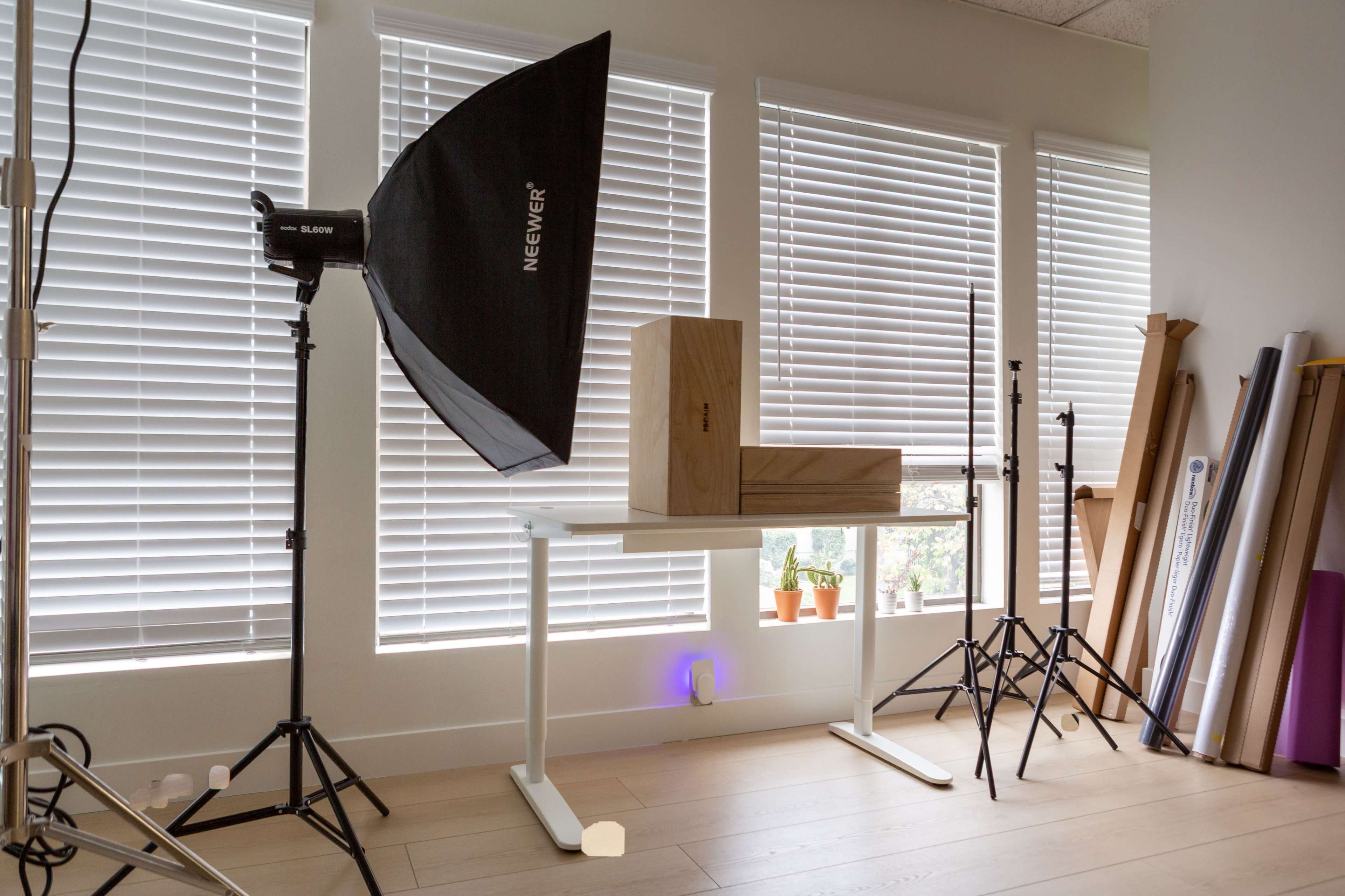 The image shows a photography setup featuring a wooden box on a white table, positioned under a large softbox light, with several light stands and rolled materials in the background.