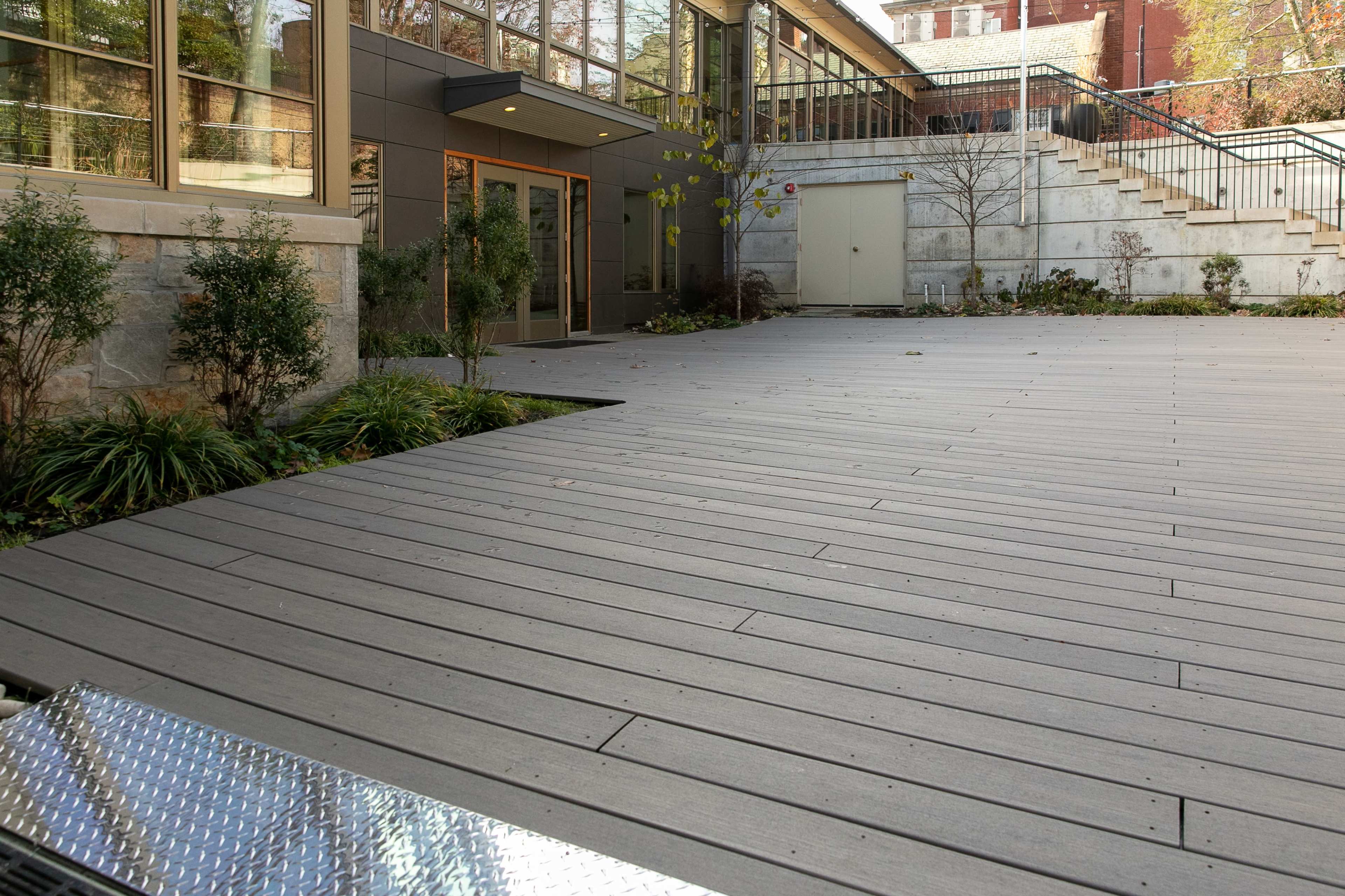 A wooden deck area adjacent to a modern building with large windows, featuring landscaped greenery and a staircase in the background.