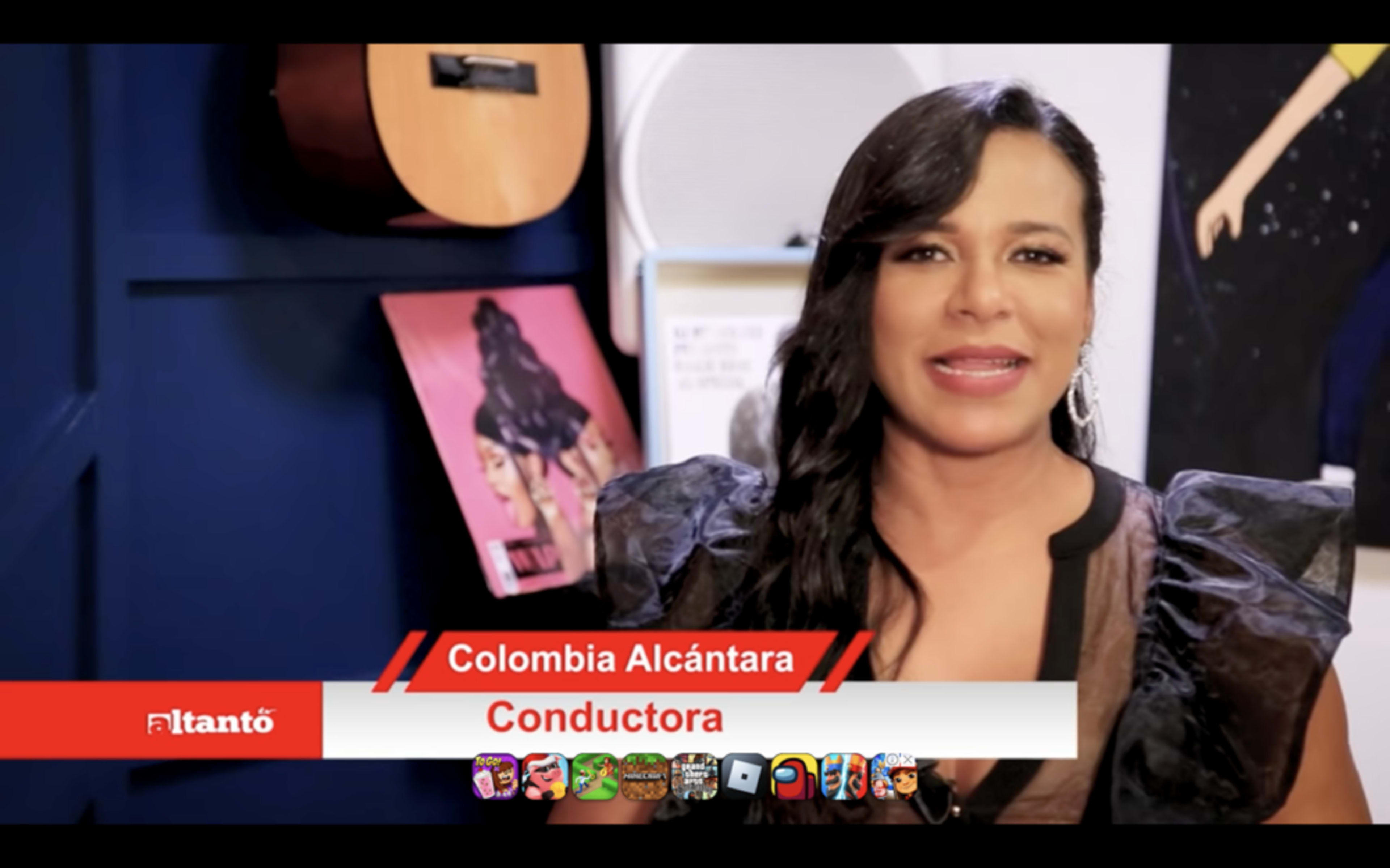 A woman stands in front of a blue wall, holding a microphone, with a guitar and a framed poster behind her.