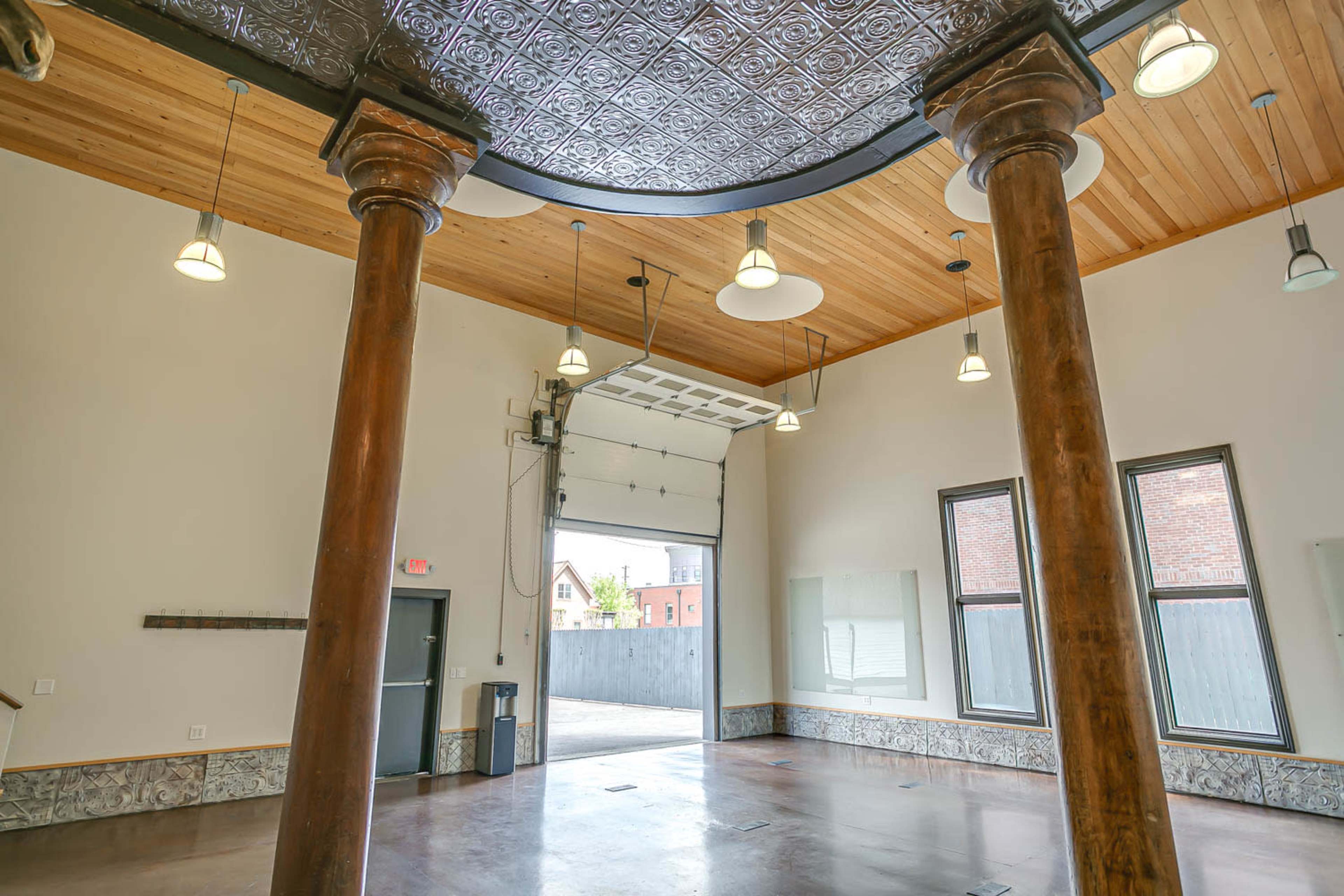The image shows an interior space featuring wooden columns, a high ceiling with circular lighting, and an open garage door leading to the outside.
