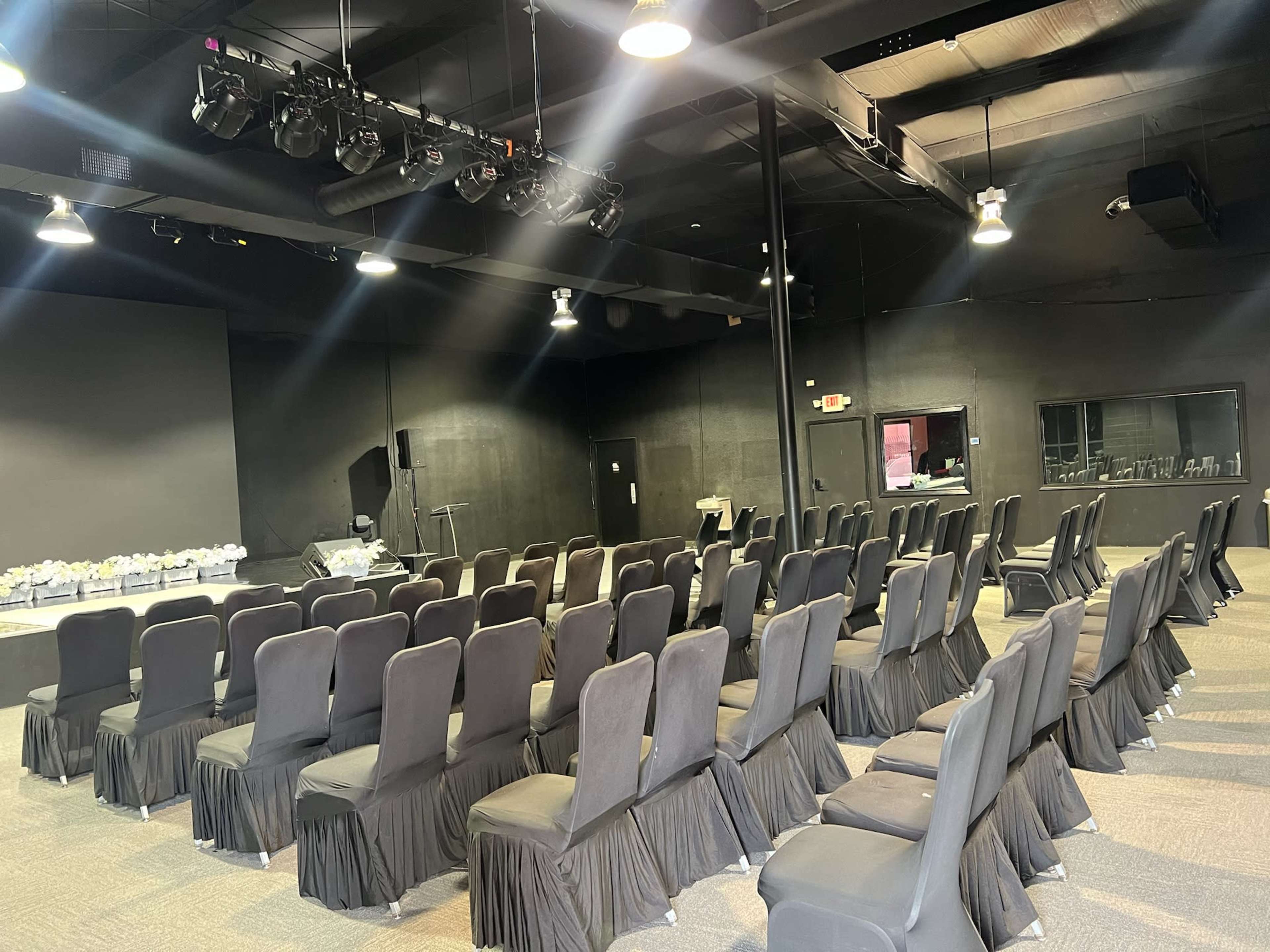 The image shows a spacious indoor venue set up with rows of black upholstered chairs facing a stage area.