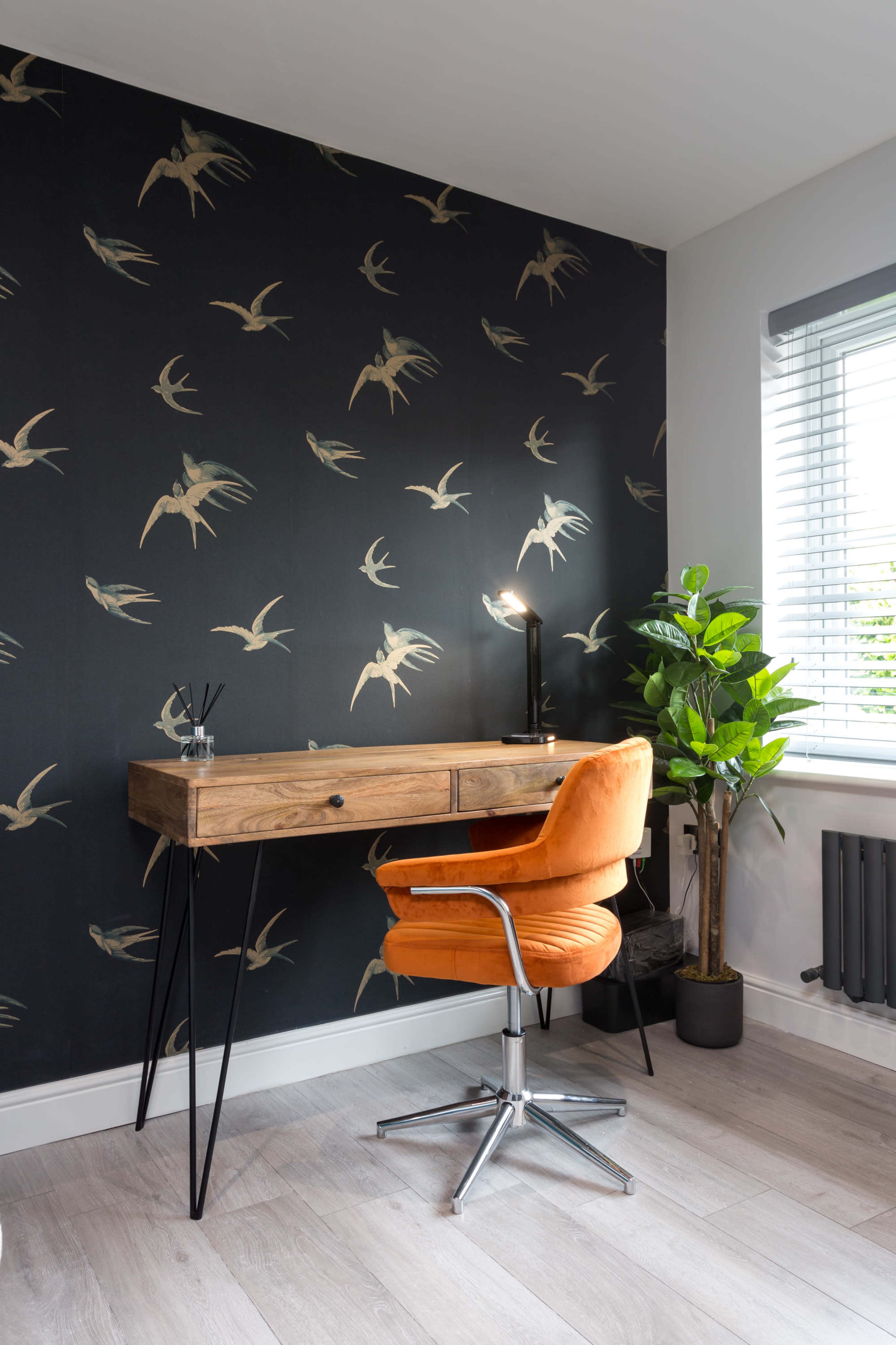 A wooden desk with a black background featuring swallow patterns is paired with an orange chair and a potted plant in a well-lit room.