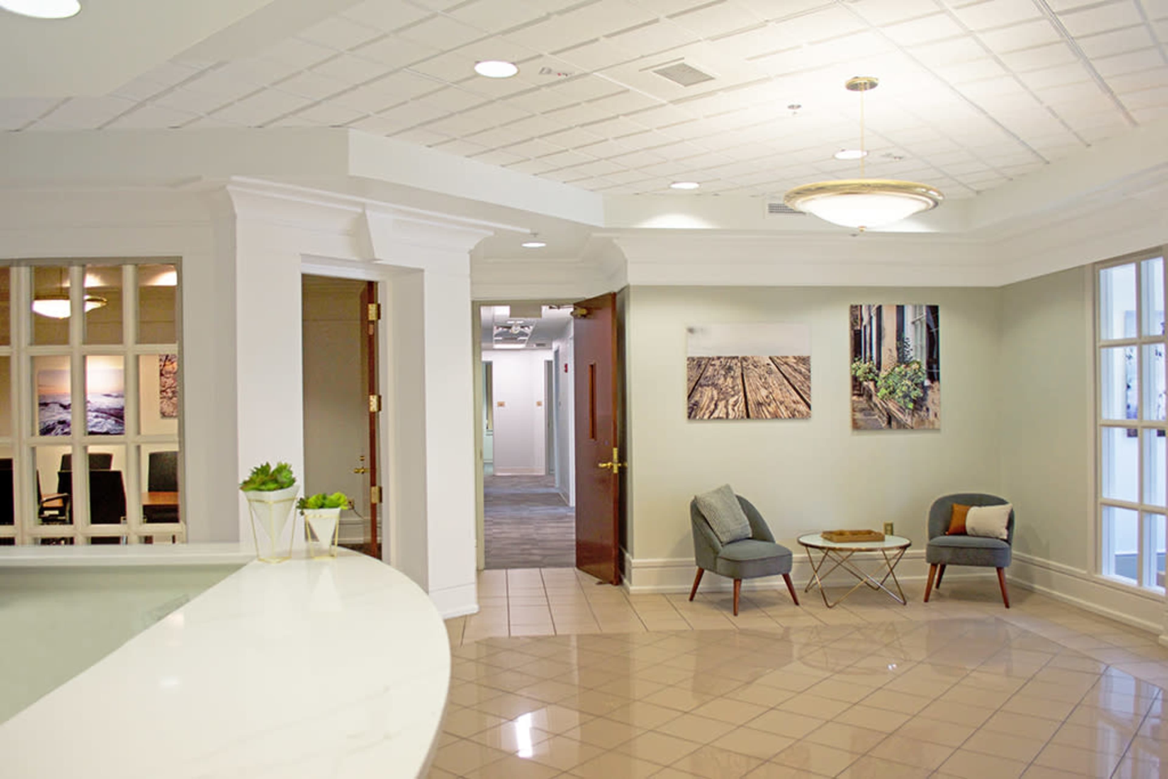 A modern reception area featuring a curved white desk, two chairs with a table, and artwork on the walls in a well-lit space.