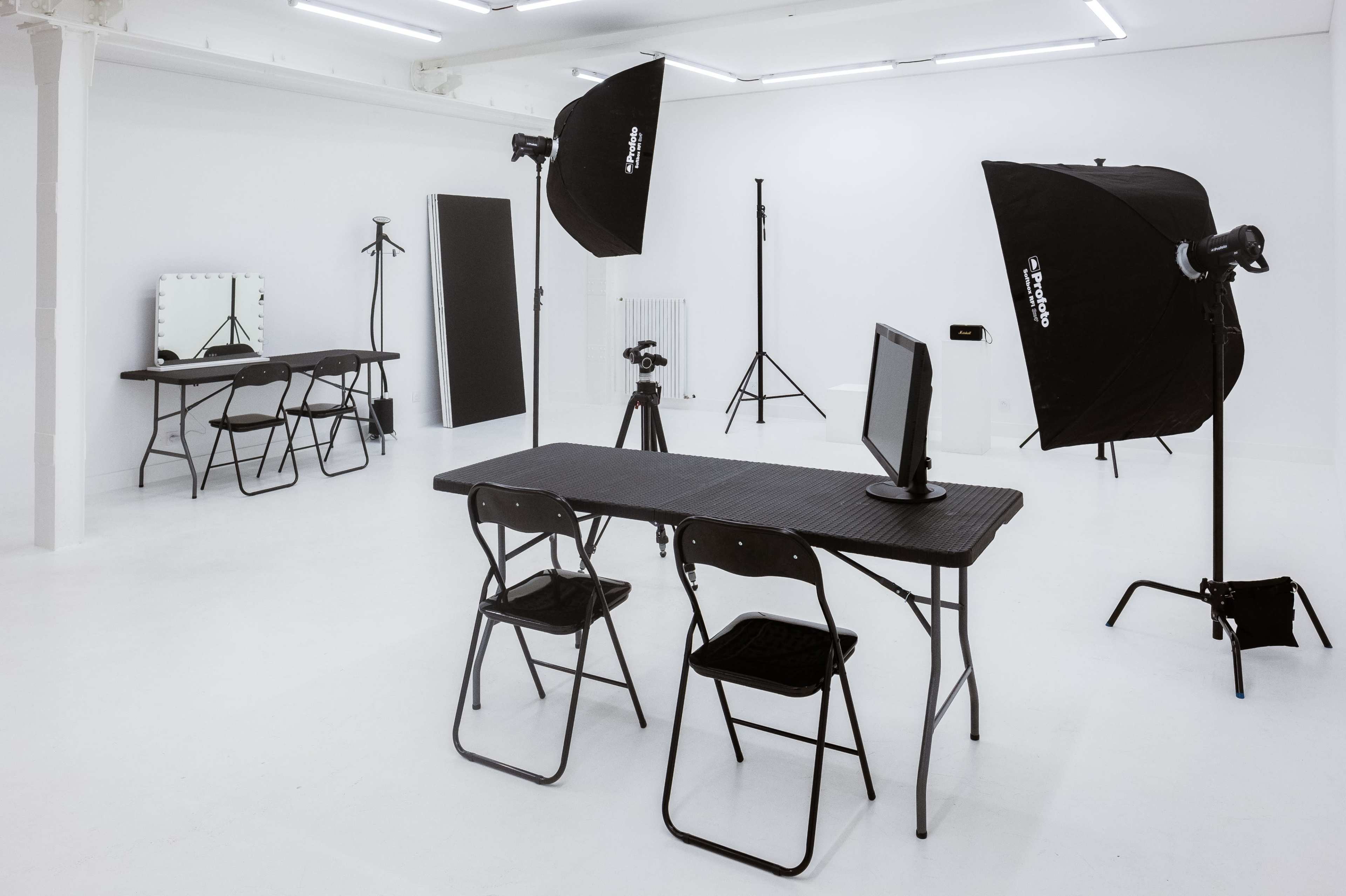 A well-lit photography studio featuring a table with chairs, a computer monitor, and various lighting equipment set up against white walls.
