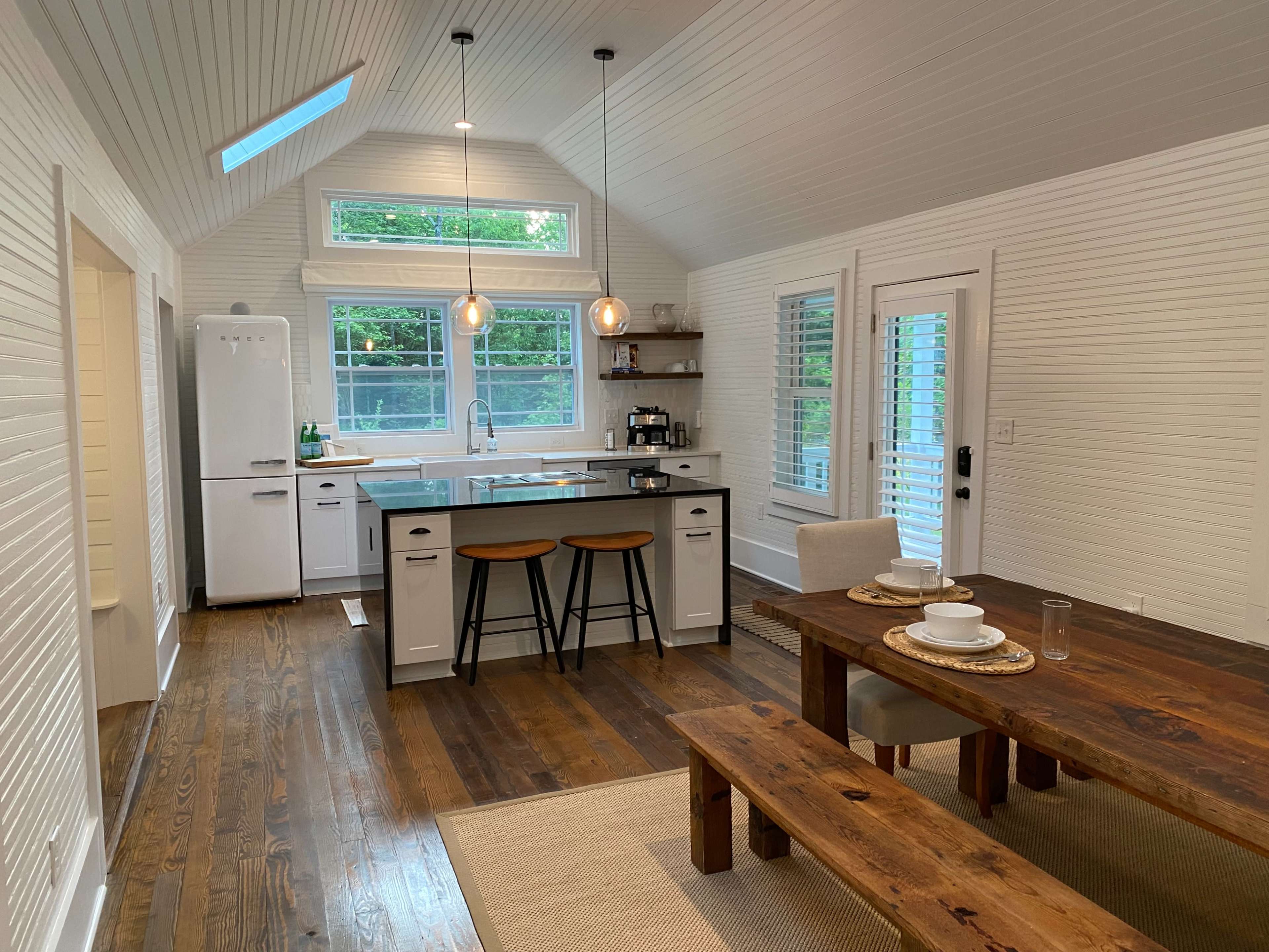 A bright, airy kitchen and dining area with a wooden dining table, two stools at a kitchen island, and large windows letting in natural light.