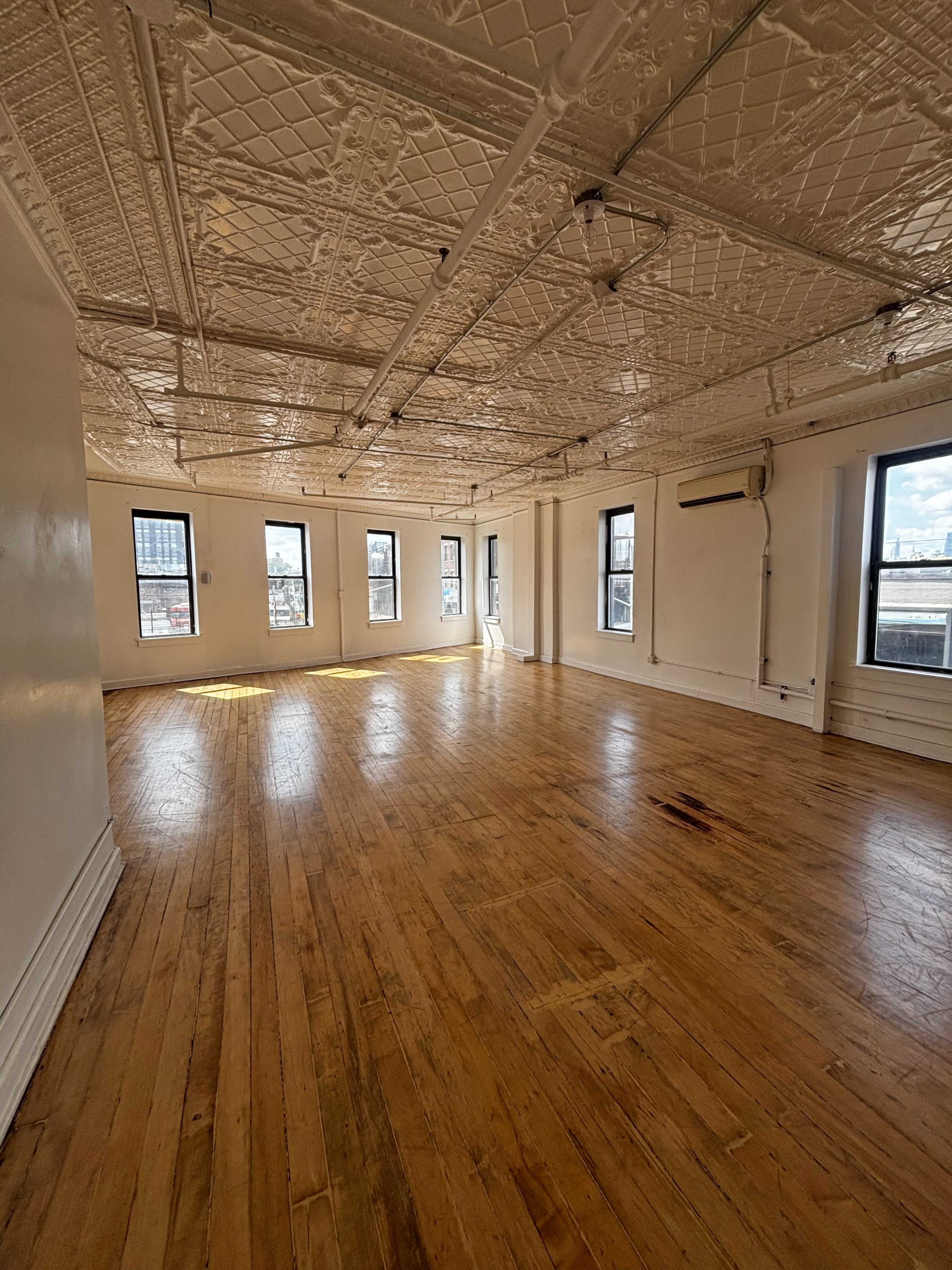 The image shows a large, empty room with wooden floors and several windows, featuring a textured ceiling.
